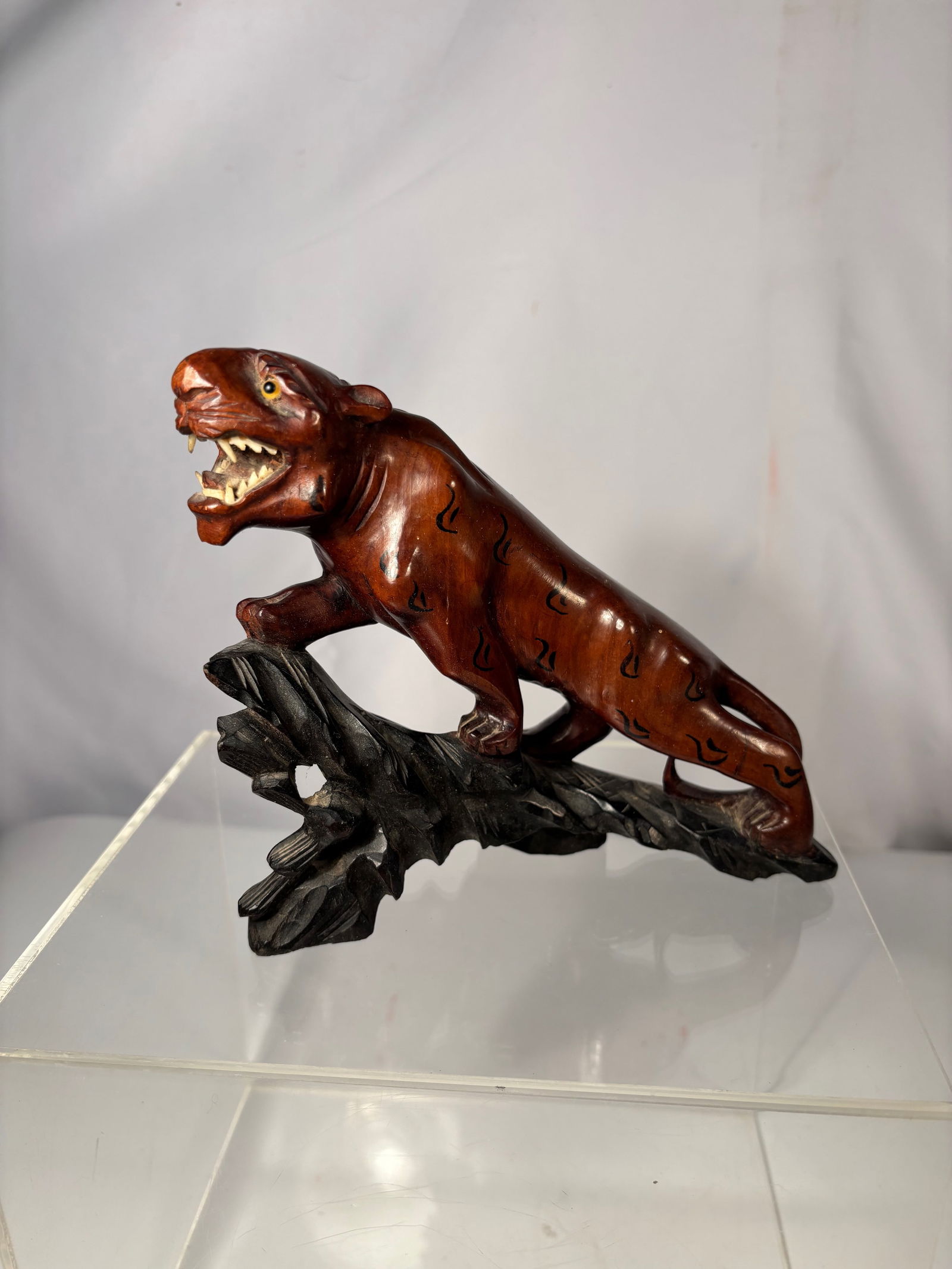 A Japanese Style Carved Tiger Figure on Wood Base: A Japanese-style carved figure depicting a tiger in a dynamic stalking pose, mounted on a carved base. The animal is rendered with expressive detail, including open jaws, incised striping, and inlaid-