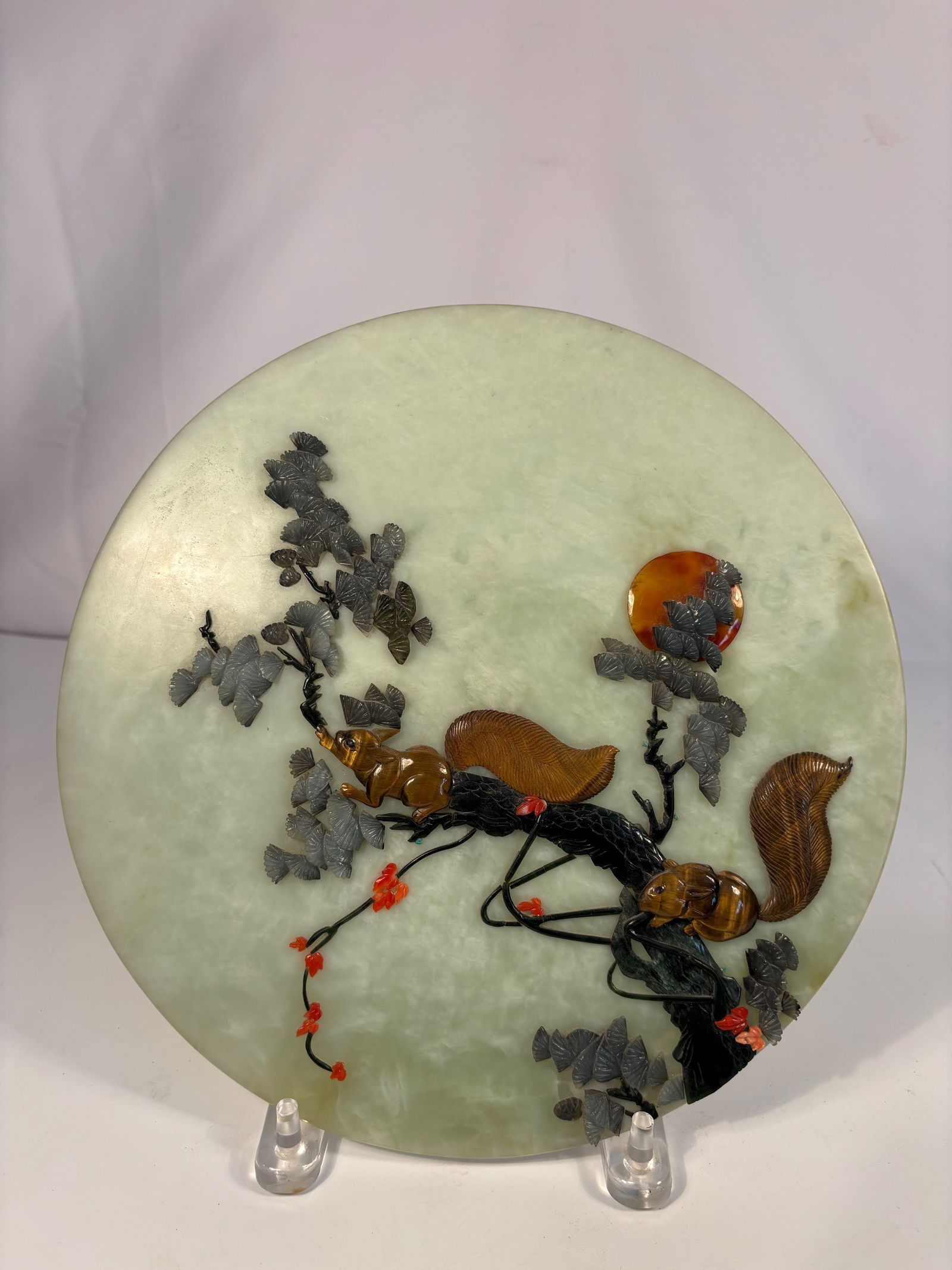 A Chinese Jade Plaque Inlaid with Coral, Agate, and Tiger’s Eye: A circular plaque of pale celadon jade, finely inlaid with coral, agate, and tiger’s eye stones to depict a lively squirrel climbing among grapevines with clusters of fruit. The squirrel’s body is