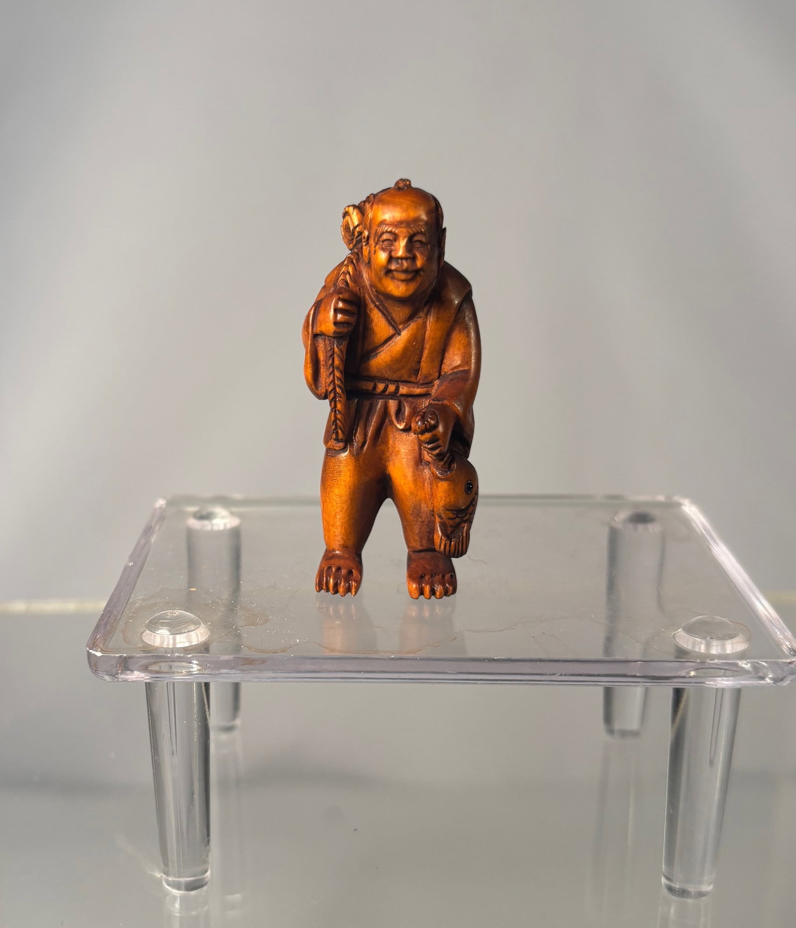 A Japanese Boxwood Katabori Netsuke – Smiling Monk Carrying a Basket, Signed Gyokusan: Meiji period (19th century), Finely carved Japanese boxwood netsuke depicting a smiling itinerant monk standing upright, carrying a woven basket on his back. The figure is rendered with expressive fac