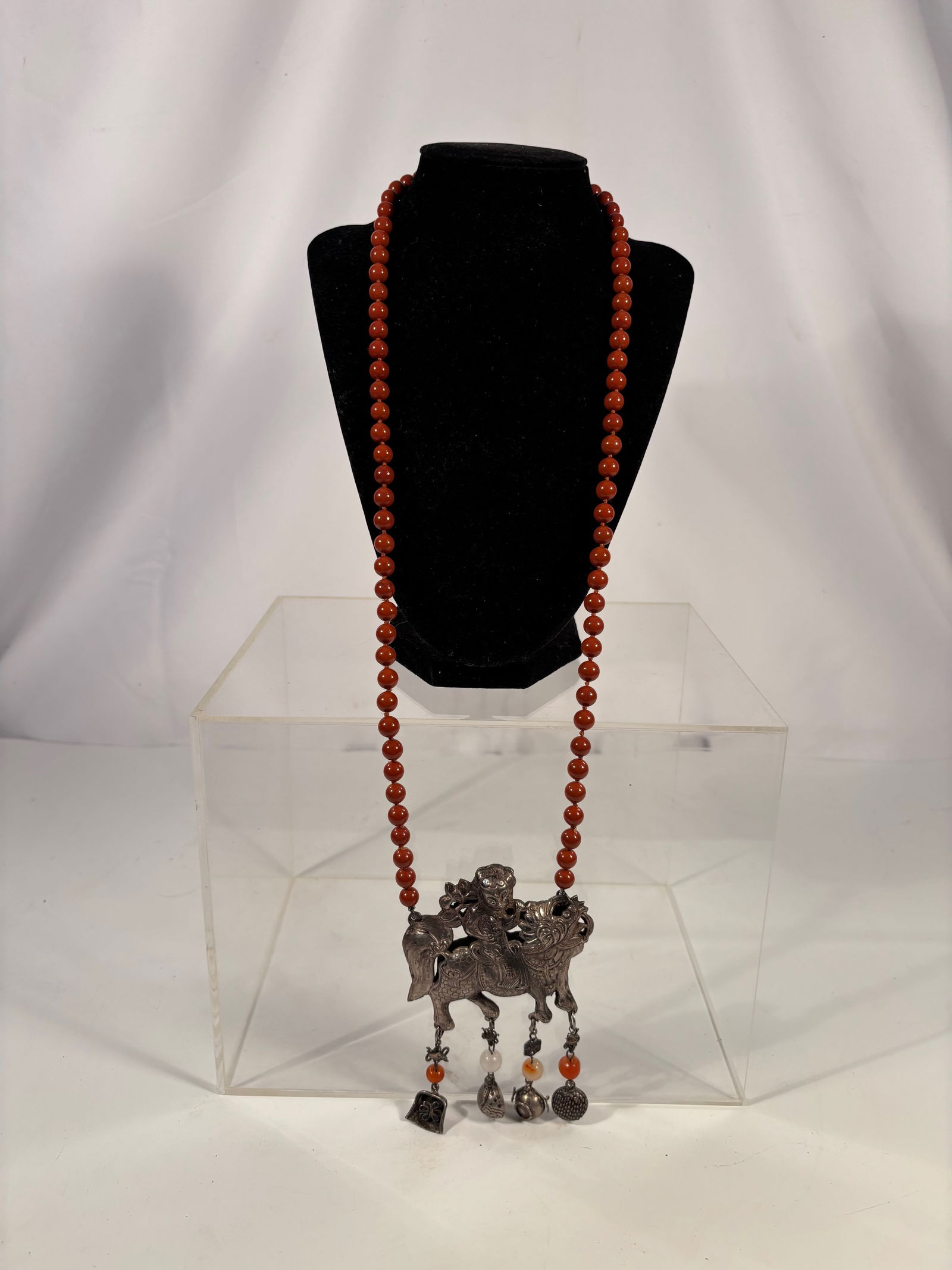 A Chinese Silver Pendant Necklace with Red Jasper Beads, Rider on Qilin: A long strand necklace composed of polished round red jasper beads, suspending a large silver pendant depicting a child or celestial rider seated upon a qilin (mythical beast). From the pendant hang m