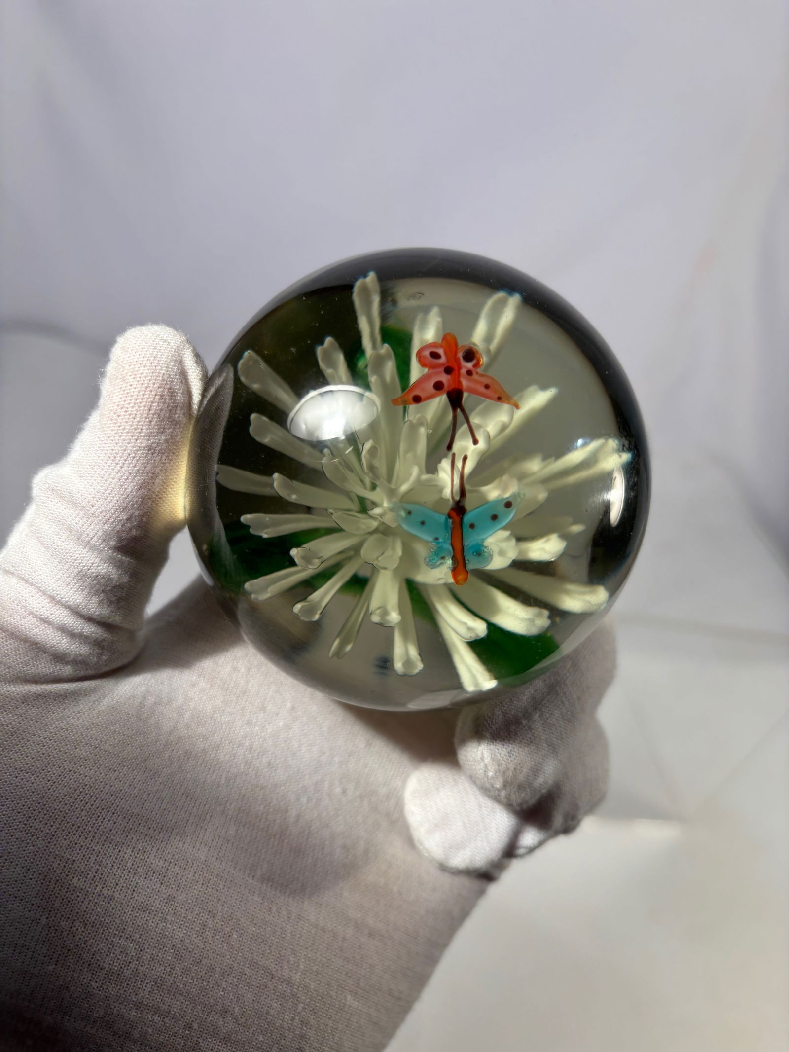 Art Glass Lampwork Floral Paperweight with Butterflies: Decorative art glass paperweight featuring a sculpted white lampwork flower rising from a green stem, set against a warm amber-toned ground. Two colorful butterflies hover above the bloom, creating a