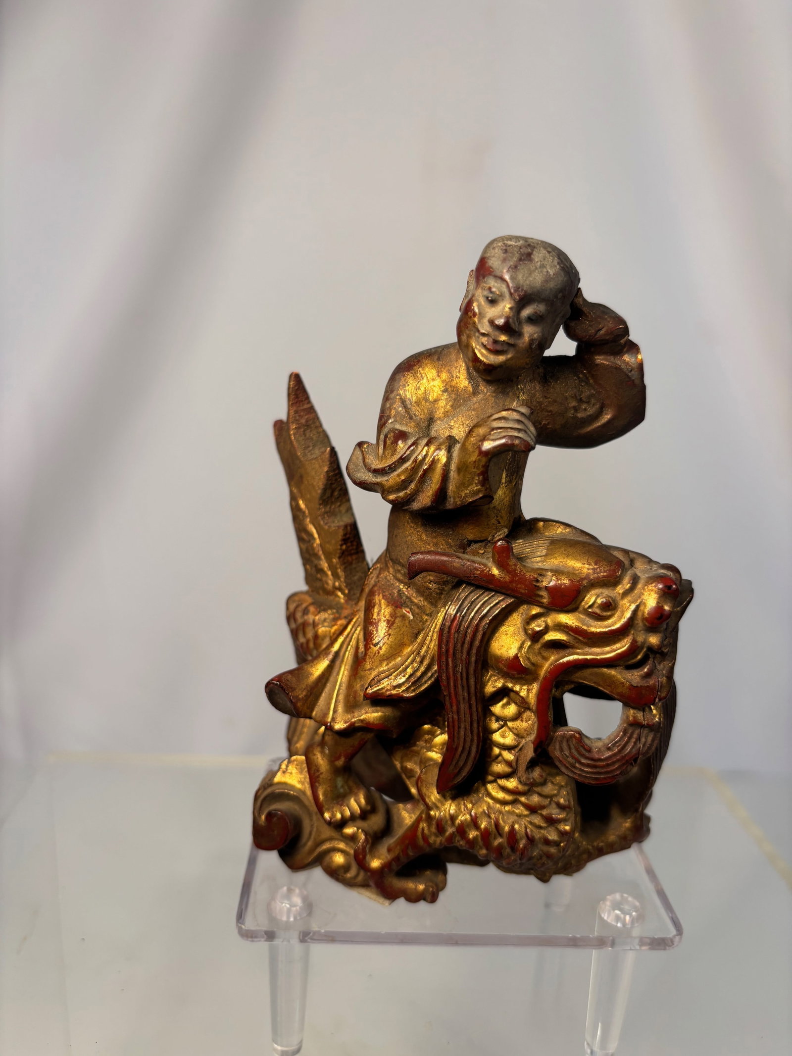 A Chinese Giltwood Figure of a Boy Riding a Dragon, Late Qing Dynasty: Qing dyansty(1644-1912); Finely carved in high relief, depicting a smiling boy seated atop a sinuous dragon, holding one hand raised to his head and the other grasping a flute or staff. The dragon’s