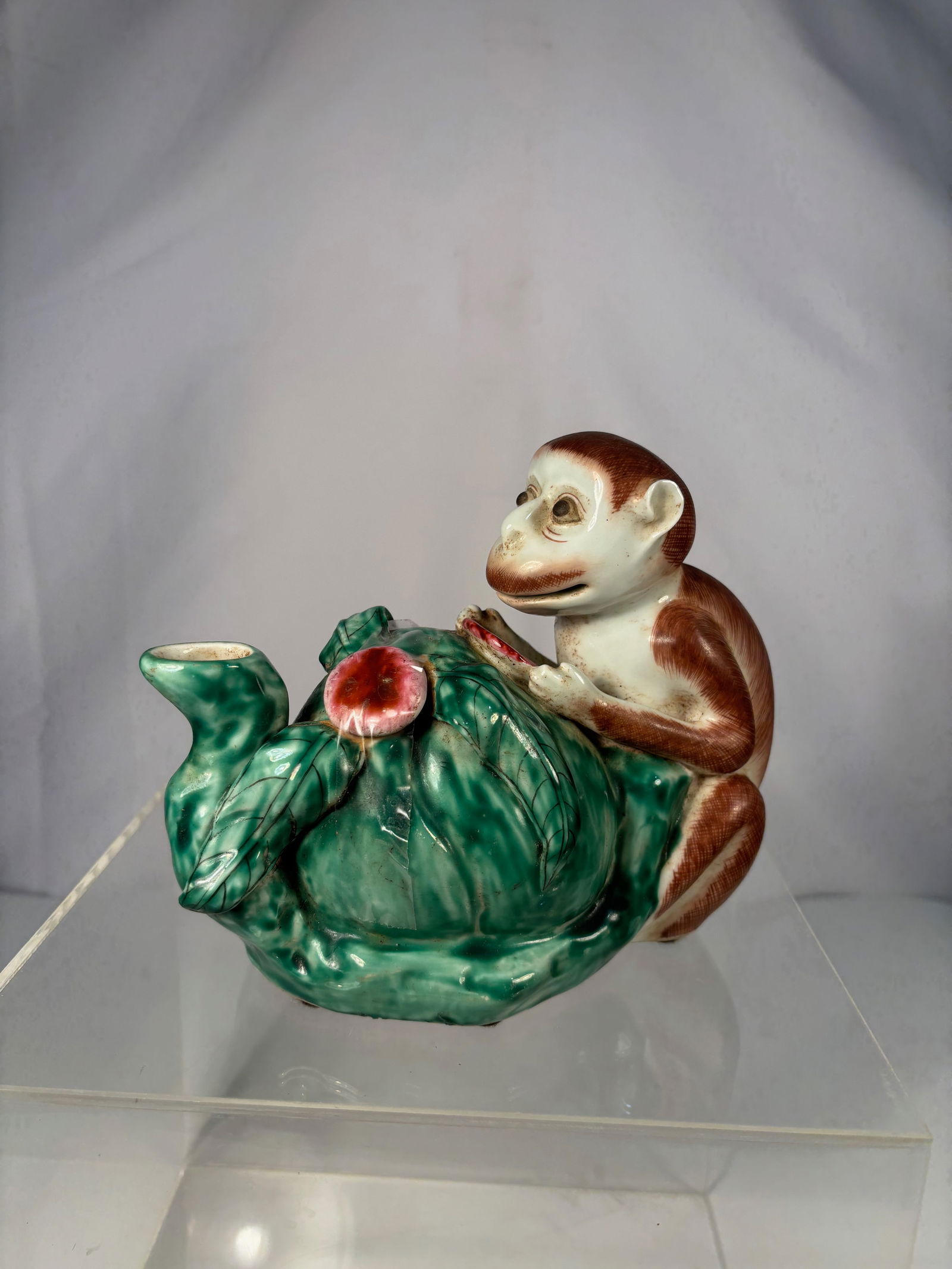 An Asian Polychrome Glazed Porcelain Teapot in the Form of a Monkey Holding a Peach: Early 20th century ; A charming Chinese porcelain figural teapot, molded as a seated monkey clutching a large peach, the body forming the vessel and spout. Decorated in vibrant sancai-style enamels of
