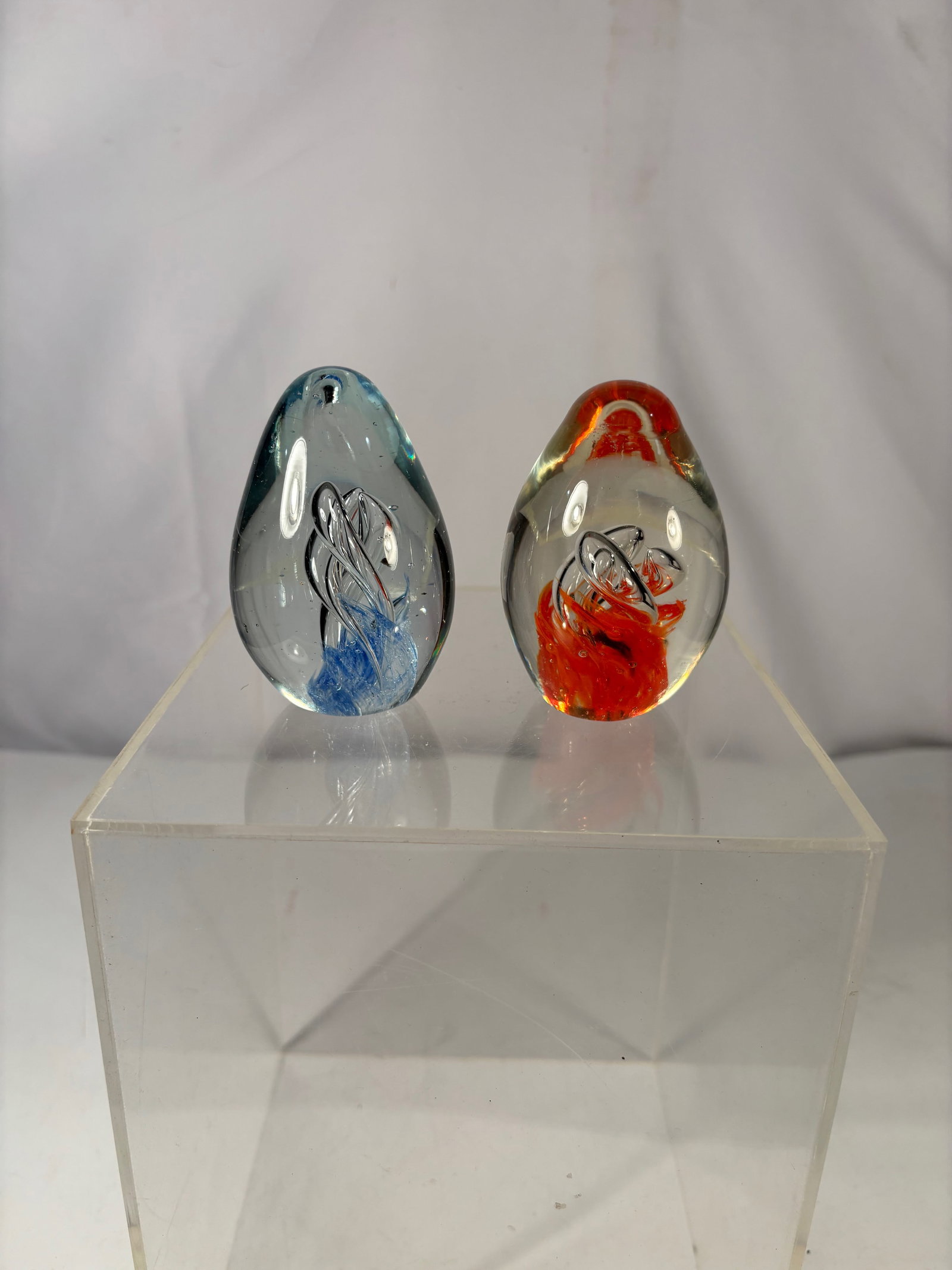 Pair of Art Glass Paperweights w/ Blue & Red Swirl Designs: A pair of decorative studio art glass paperweights in freeform teardrop shapes, each featuring controlled internal ribbon-like swirl inclusions. One example with blue tones, the other with red/orange