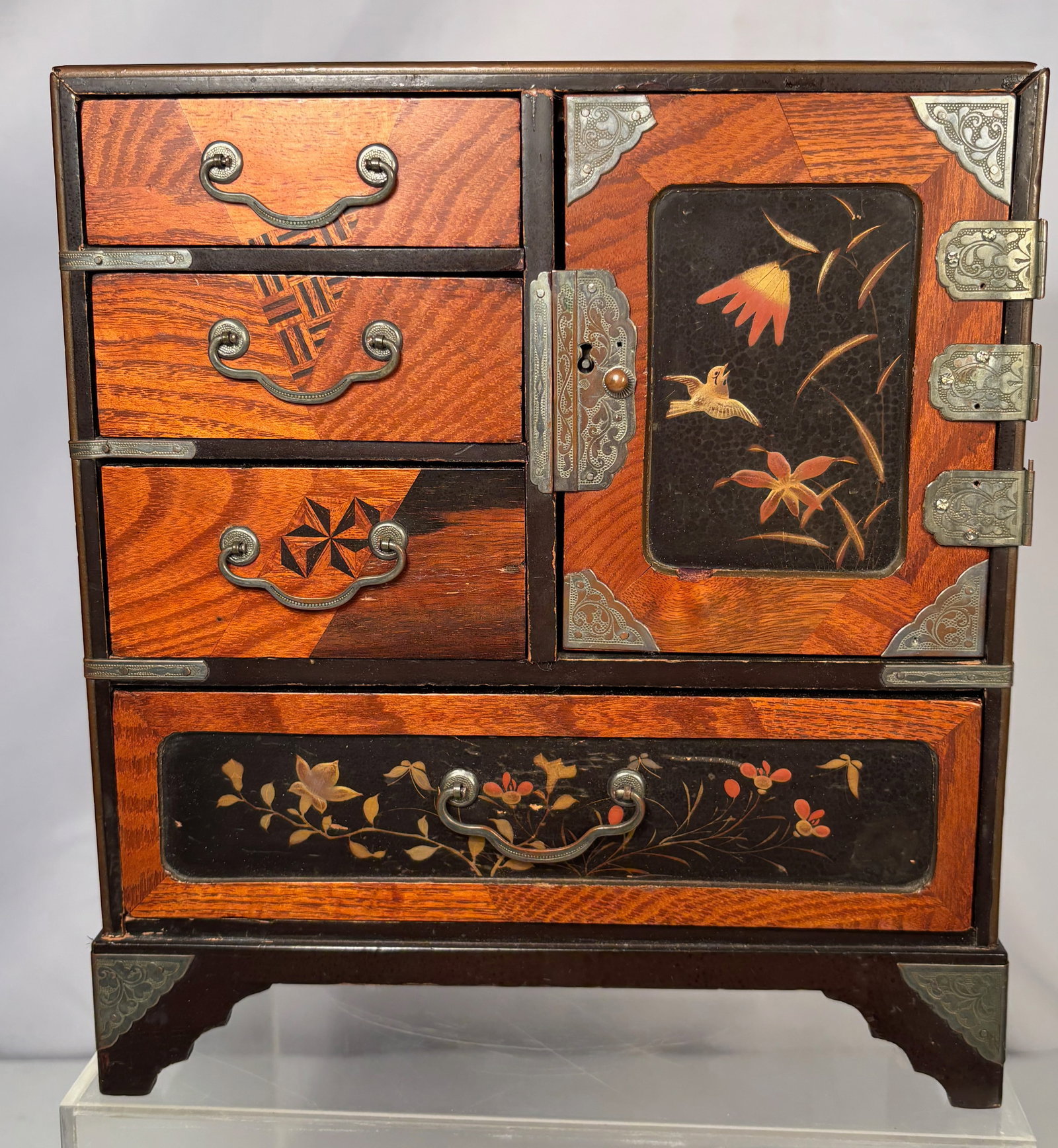 A Japanese small Tansu Style Jewelry Chest with Floral Decoration: A Japanese miniature tansu style cabinet, having three small drawers, a side cupboard door, and a lower drawer, with decorative metal mounts throughout. The case is decorated with floral and bird moti