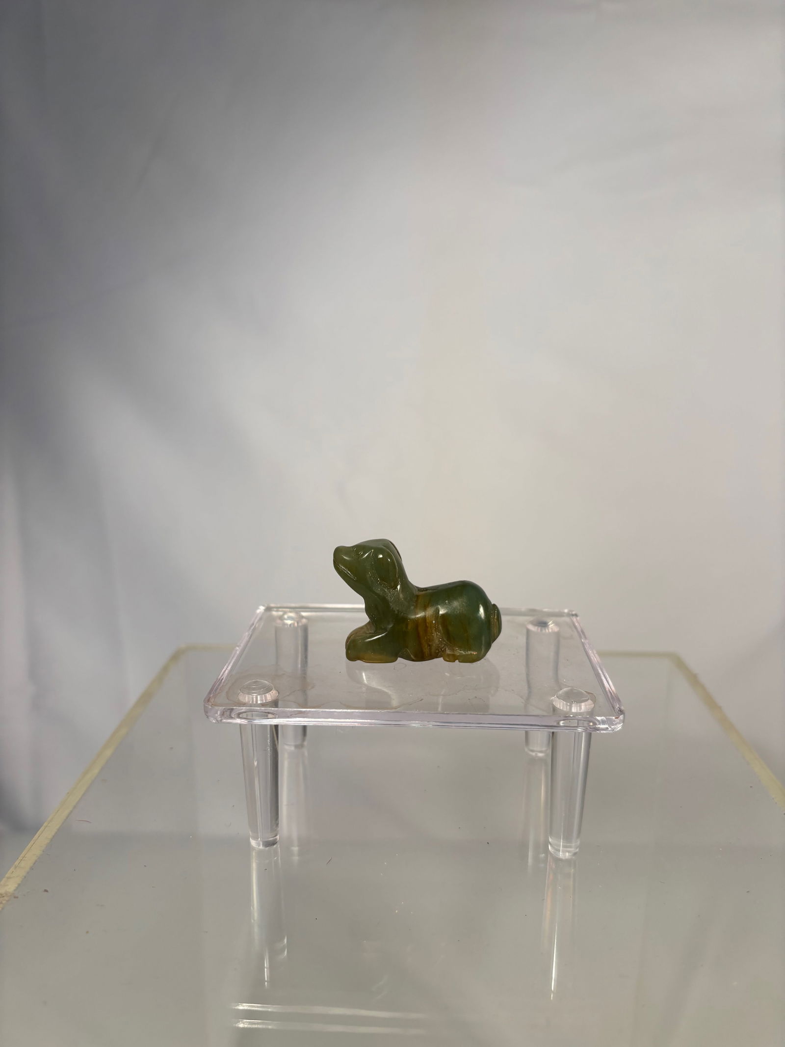 Chinese Carved Green Jadeite Dog: A delicately carved Chinese jadeite figure of a dog, presented in a crouching position with stylized detailing of ears, limbs, and eyes. The piece is crafted from deep green translucent jadeite, polis