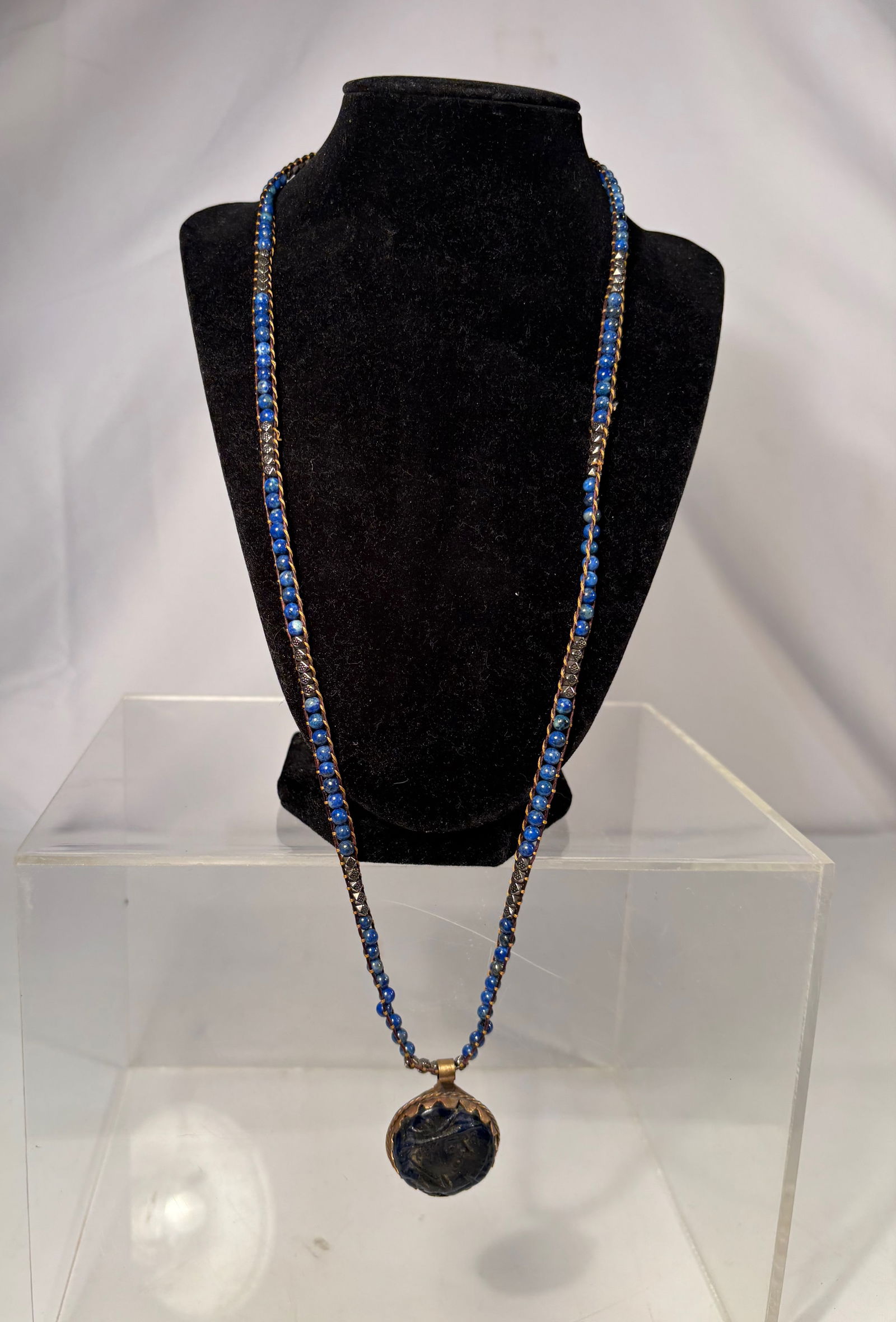 A Vintage Lapis Lazuli Beaded Necklace with Carved Hardstone Pendant: A vintage necklace composed of small lapis lazuli beads, arranged in multiple strands with dark metal spacer elements. The necklace is centered by a hand-carved hardstone pendant, dark green to black