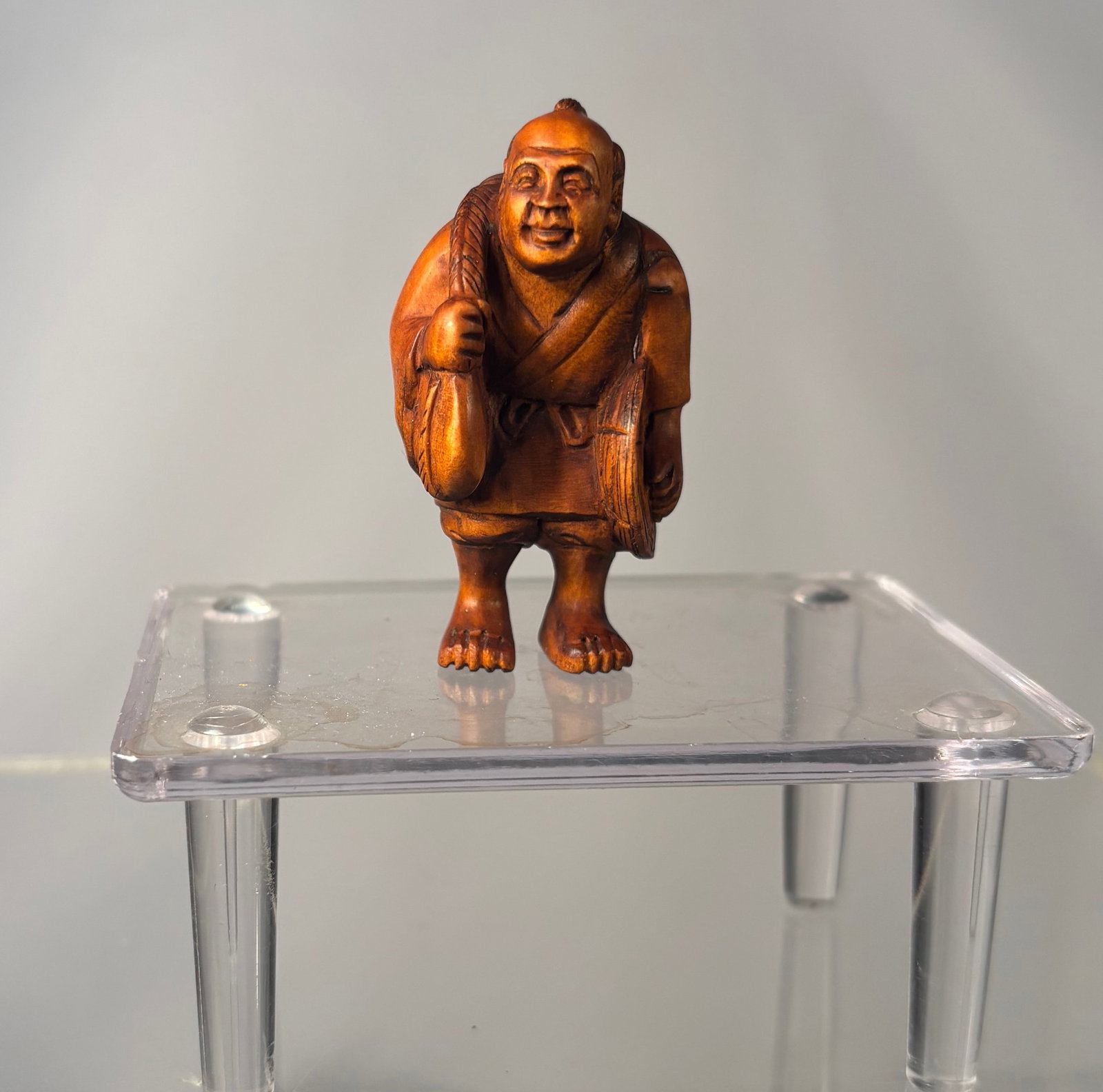 A Japanese Boxwood Netsuke – Smiling Itinerant Monk with Human Face Carved on His Back, Signed: Meiji period (19th century), Finely carved Japanese boxwood netsuke depicti ng a smiling itinerant monk standing upright, his robe rendered with fluid, natural folds. Notably, a small human face is in