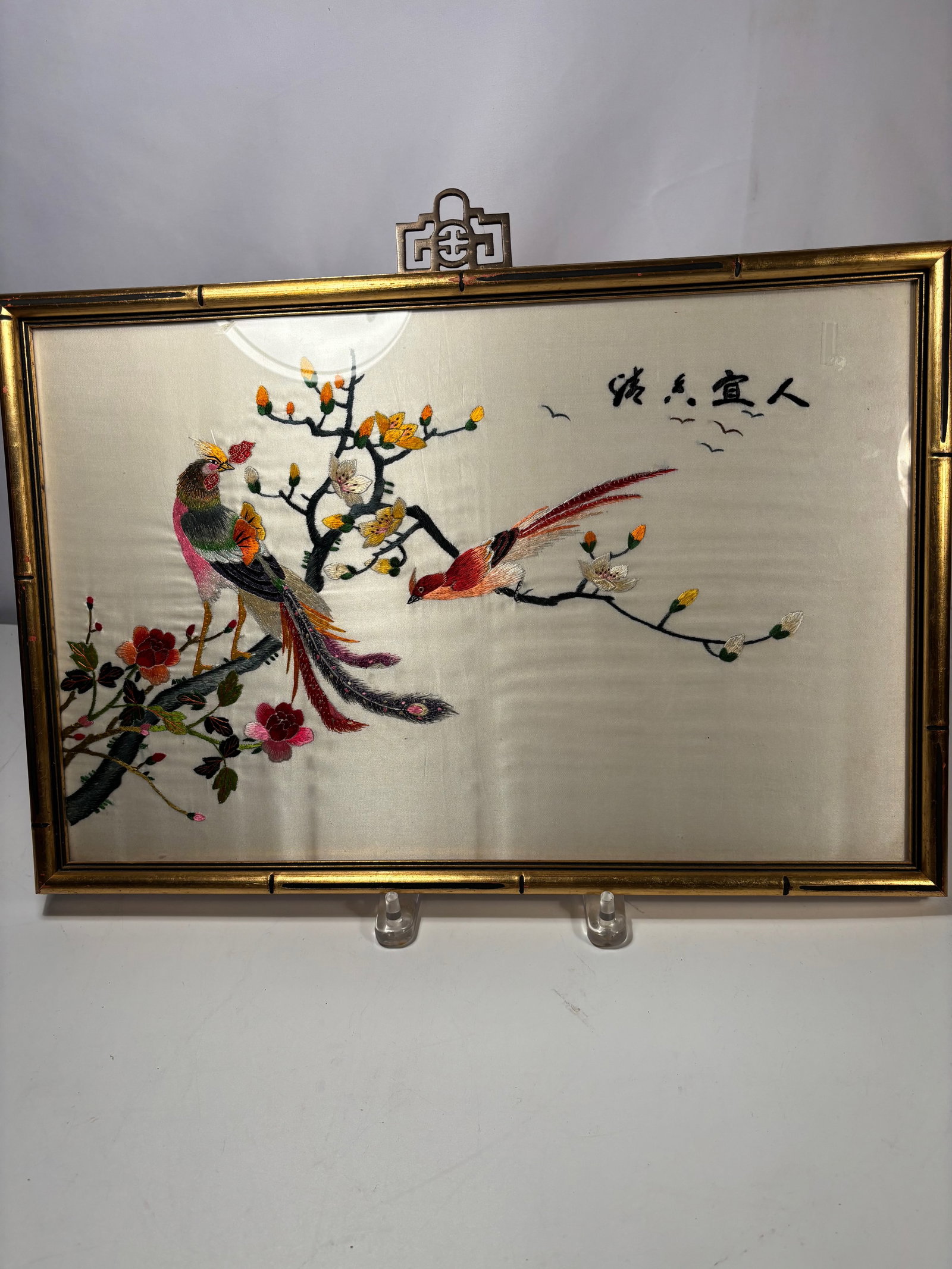 A Chinese Silk Embroidered Panel with Birds and Pine Tree, Framed: A finely executed Chinese silk embroidered panel depicting a colorful bird perched on a flowering branch, with a second bird in flight nearby. The scene is rendered in vibrant silk threads with detail