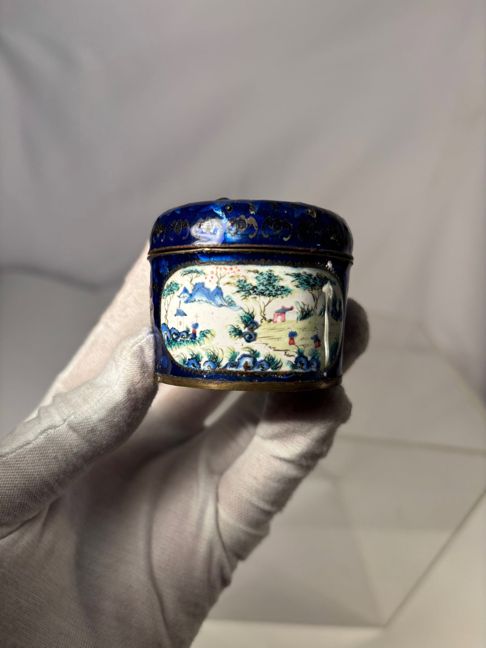 A Chinese Blue Enamel Lidded Snuff Box with Figural Landscape: Qing dynasty(1644-1912); A Chinese enameled metal snuff box of cylindrical form, decorated on the exterior in deep cobalt-blue enamel and centered with a painted famille rose–style landscape panel d