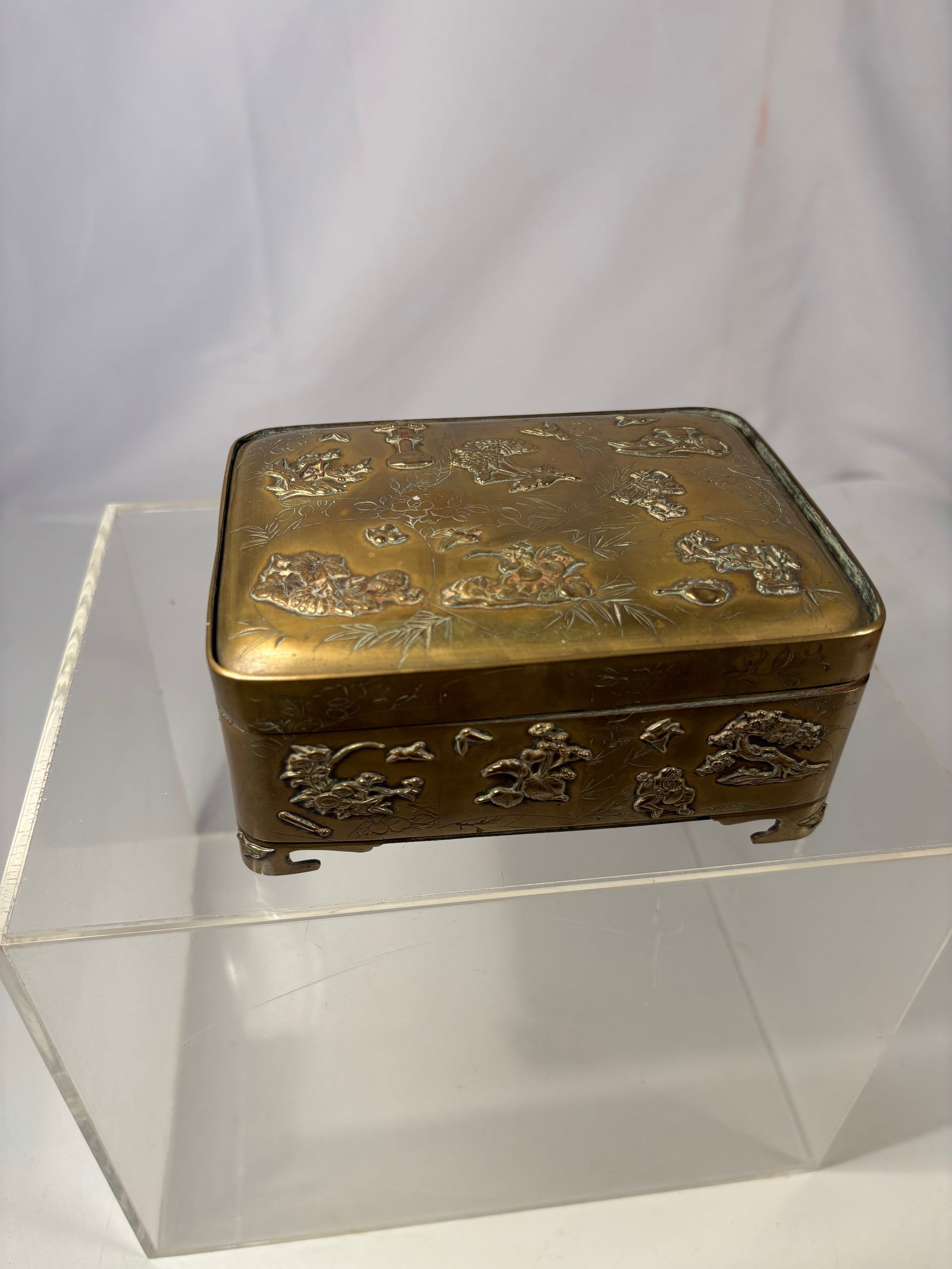 A Japanese Figural Brass Relief Box (MEIJI PERIOD 1868-1912): Japanese Meiji style rectangular brass box decorated with applied relief panels depicting figural, floral, bird, and traditional symbolic motifs. The lid features chrysanthemum and treasure objects ar