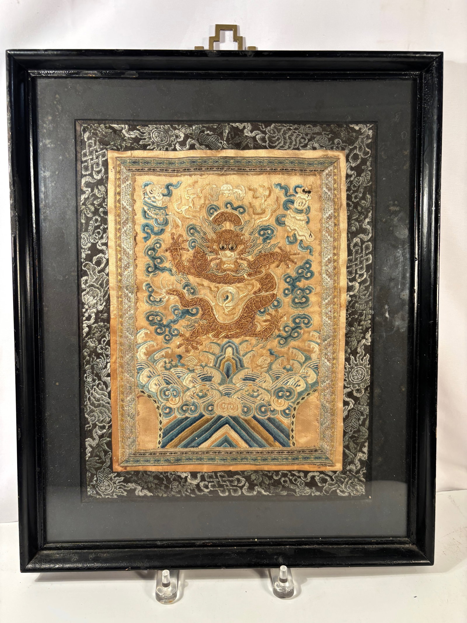 A Chinese Gold Thread Embroidered Dragon Panel, Qing Dynasty: Qing Dynasty (1644–1911); Chinese silk embroidery panel depicting a front-facing dragon executed in gold metallic thread against a ground of stylized cloud scrolls and wave (jiangya) motifs. The dra