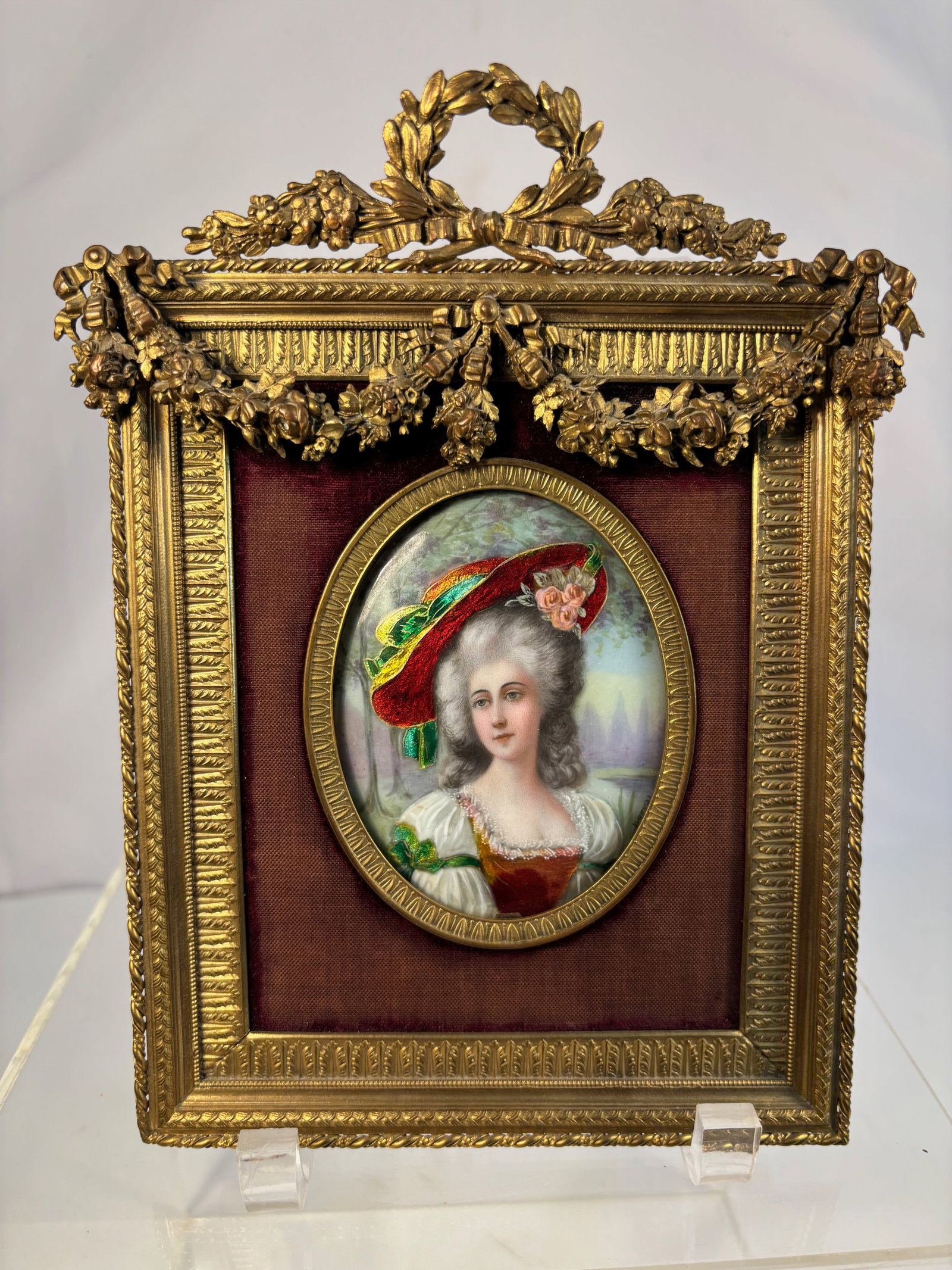 A French Gilt Bronze Framed Enamel Portrait Miniature of a Lady, Late 19th Century: Finely hand-painted on enamel, depicting an elegant lady in 18th-century attire with a crimson hat trimmed in green ribbon and pink rose, her powdered hair softly modeled and her expression serene. Th