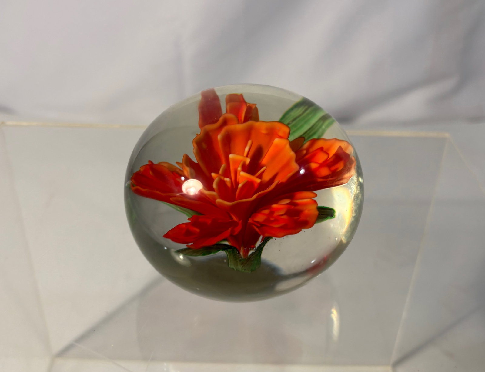 A Studio Art Glass Lampwork Floral Paperweight with Orange Flower: Studio art glass paperweight featuring a hand-formed lampwork flower in vivid orange tones with green leaves, encased within a clear spherical glass dome. The sculptural floral motif shows layered pet