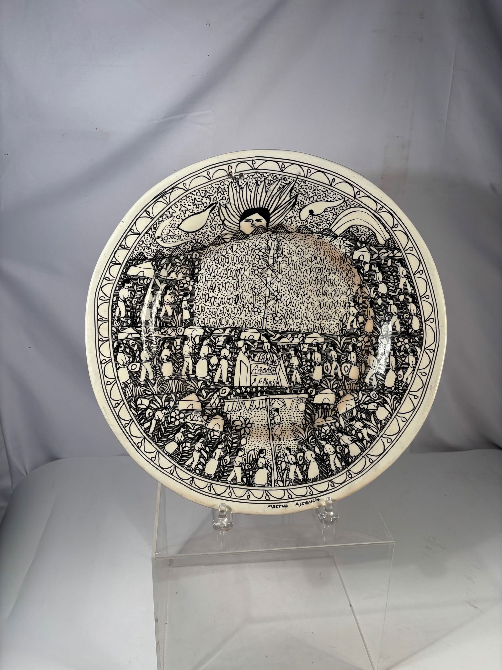 A Large Mexican Folk Art Pottery Plate by Martha Ascencio: A Mexican folk art earthenware plate decorated in black slip with a detailed narrative village scene depicting figures within a patterned landscape. Signed “Martha Ascencio” along the rim. Han