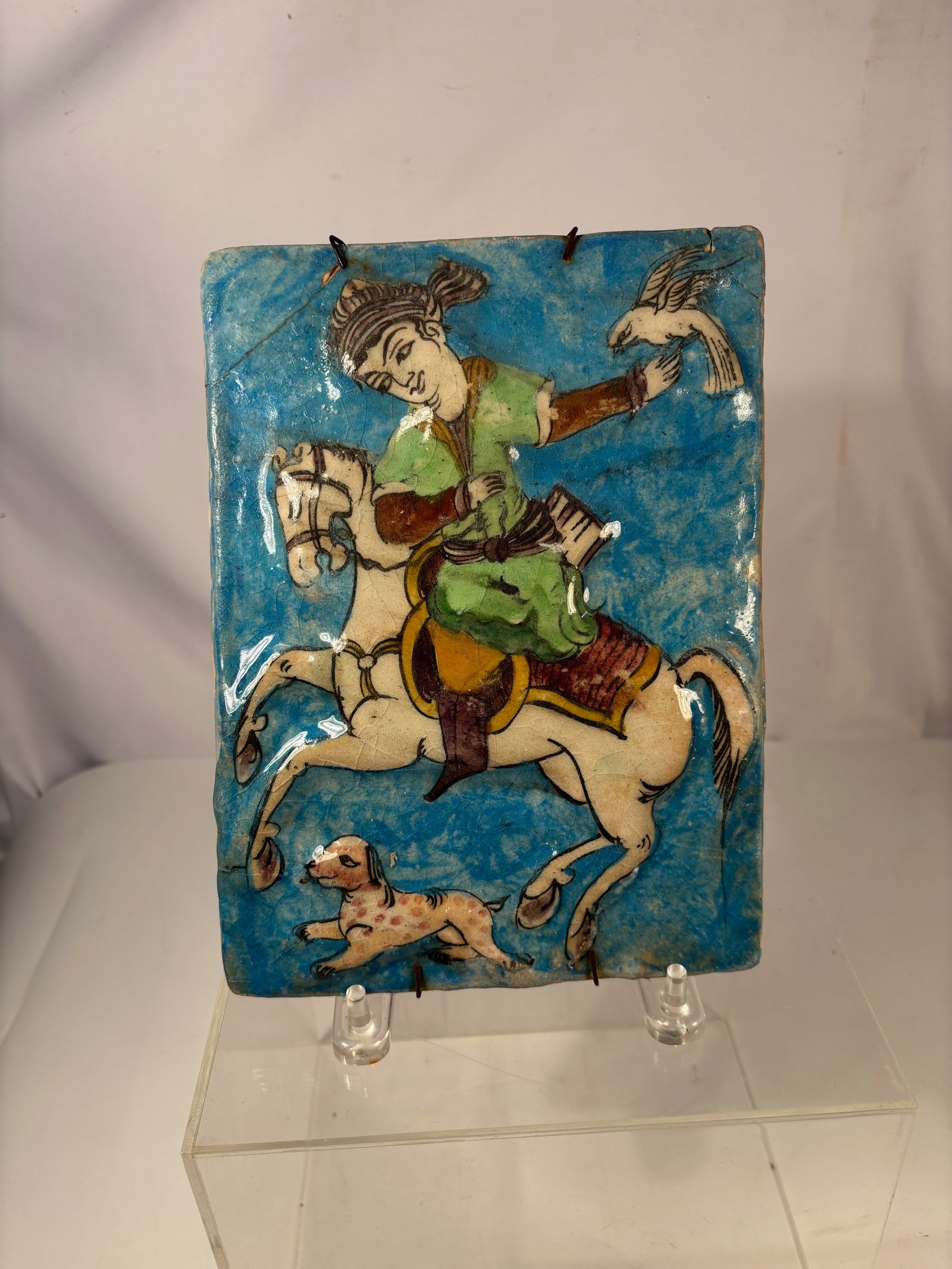 A Persian Qajar Style Glazed Ceramic Tile, 19th Century Style: A Persian glazed ceramic tile depicting a mounted falconer holding a bird of prey, accompanied by a hunting dog below. The figure is rendered in polychrome enamels against a turquoise blue ground, cha