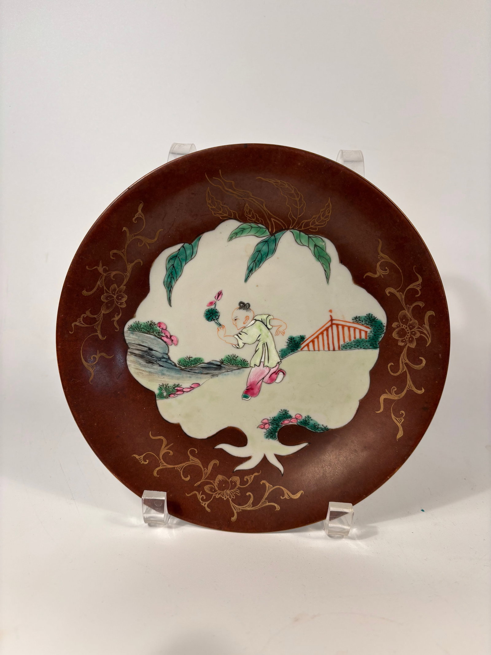 A Chinese famille rose porcelain plate with "Qianlong Mark": Qianlong period (1736-1795); A child in a garden setting holding a flower, surrounded by a brown-ground enamel border with gilt floral scrollwork — a theme symbolic of innocence, prosperity, and fil