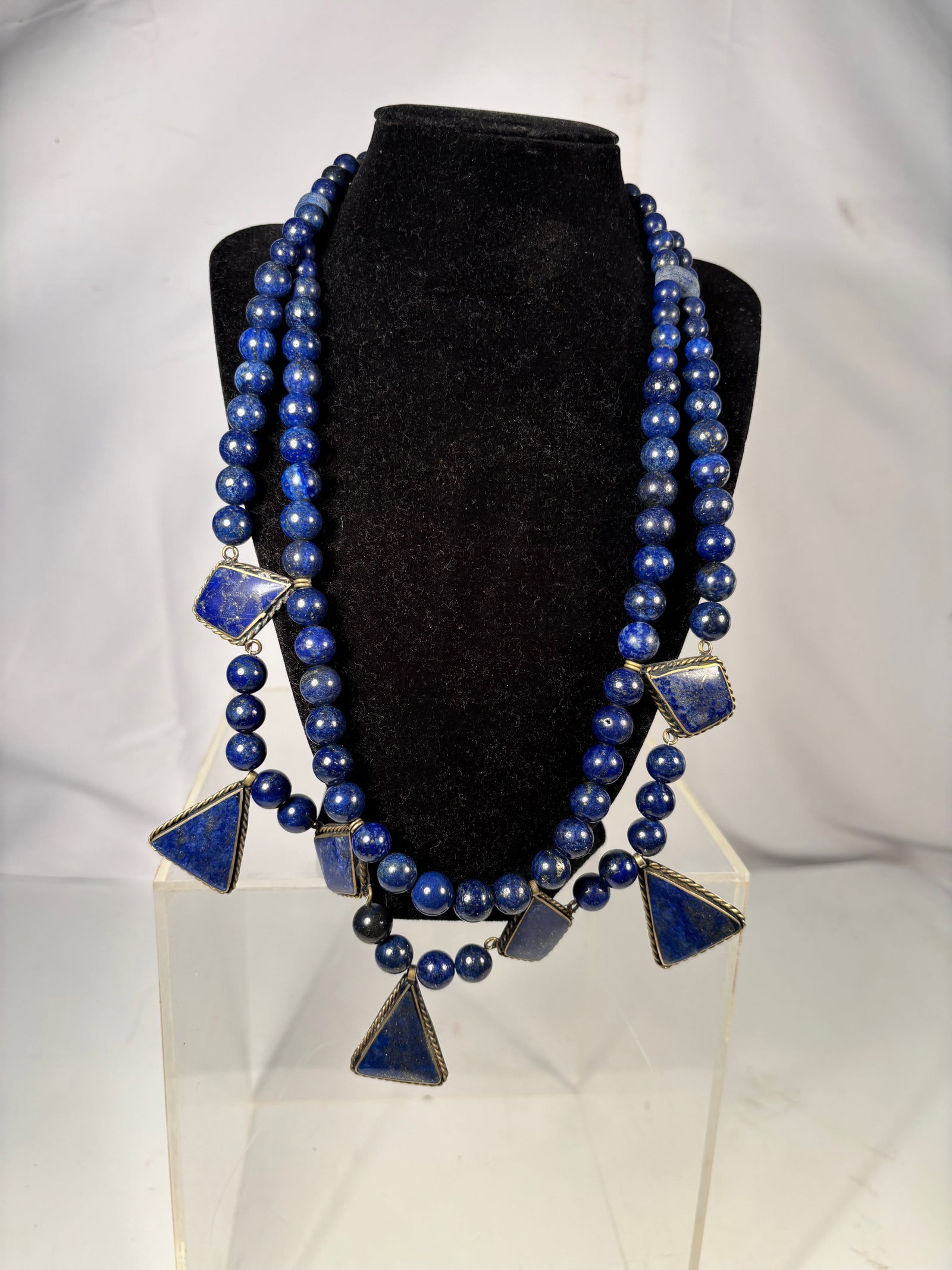 A Vintage Lapis Lazuli Bead Necklace with Geometric Pendant Drops: Vintage double strand necklace composed of polished lapis lazuli beads suspending geometric triangular pendant drops. The pendants set in metal frames with wire mounts. Rich deep blue stones with natu