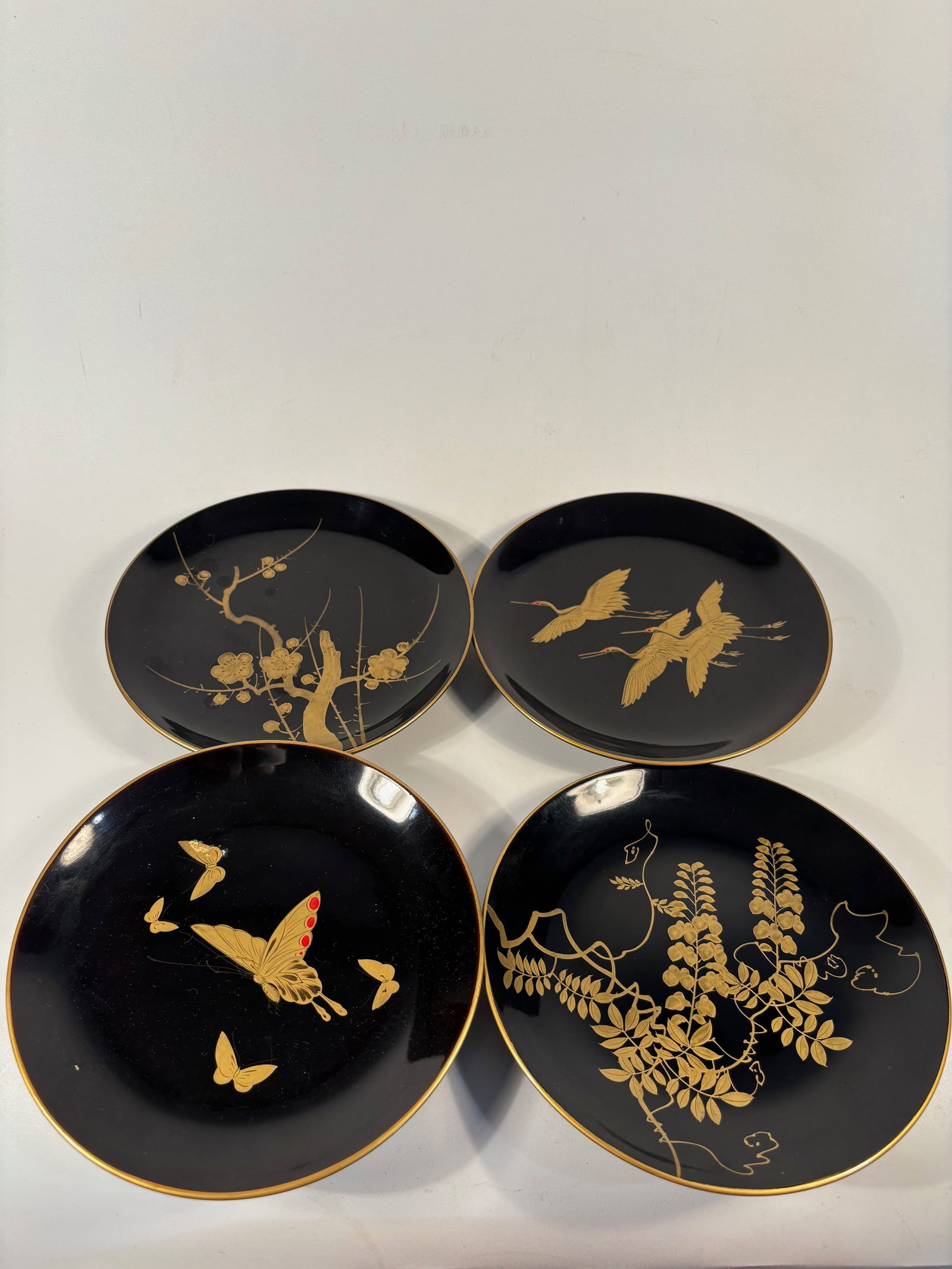 Set of 4 Japanese Makie Lacquer plates: Each plate finely decorated in gold maki-e on black lacquer with traditional Japanese motifs including plum blossoms, cranes in flight, butterflies, and flowering vines. The designs are delicately ren