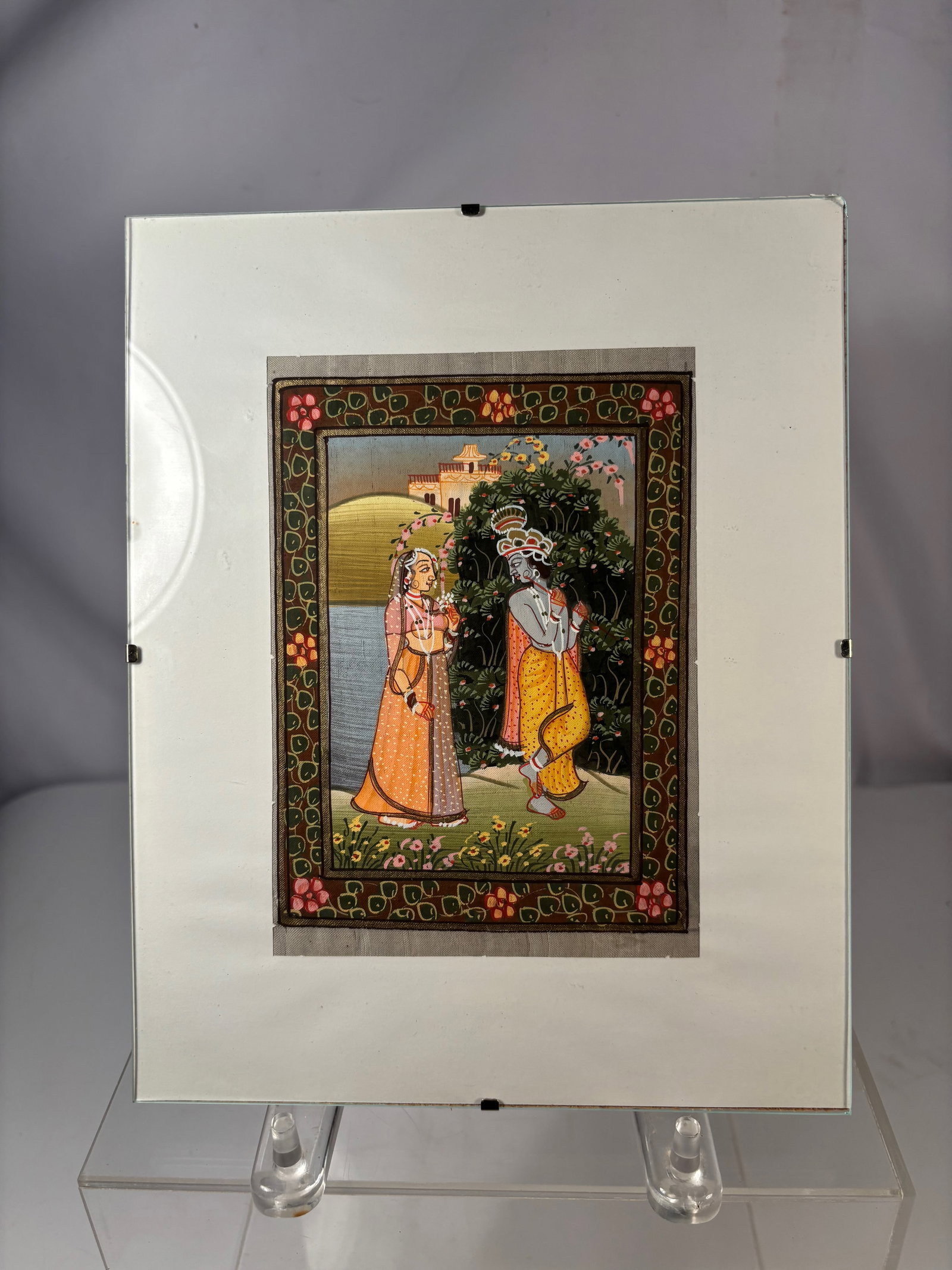 An Indian Miniature Painting of Radha and Krishna in Garden, Framed Under Glass: Hand-painted Indian miniature painting depicting Radha and Krishna standing in a lush garden landscape. Krishna is shown playing the flute while Radha stands beside him, surrounded by flowering plants
