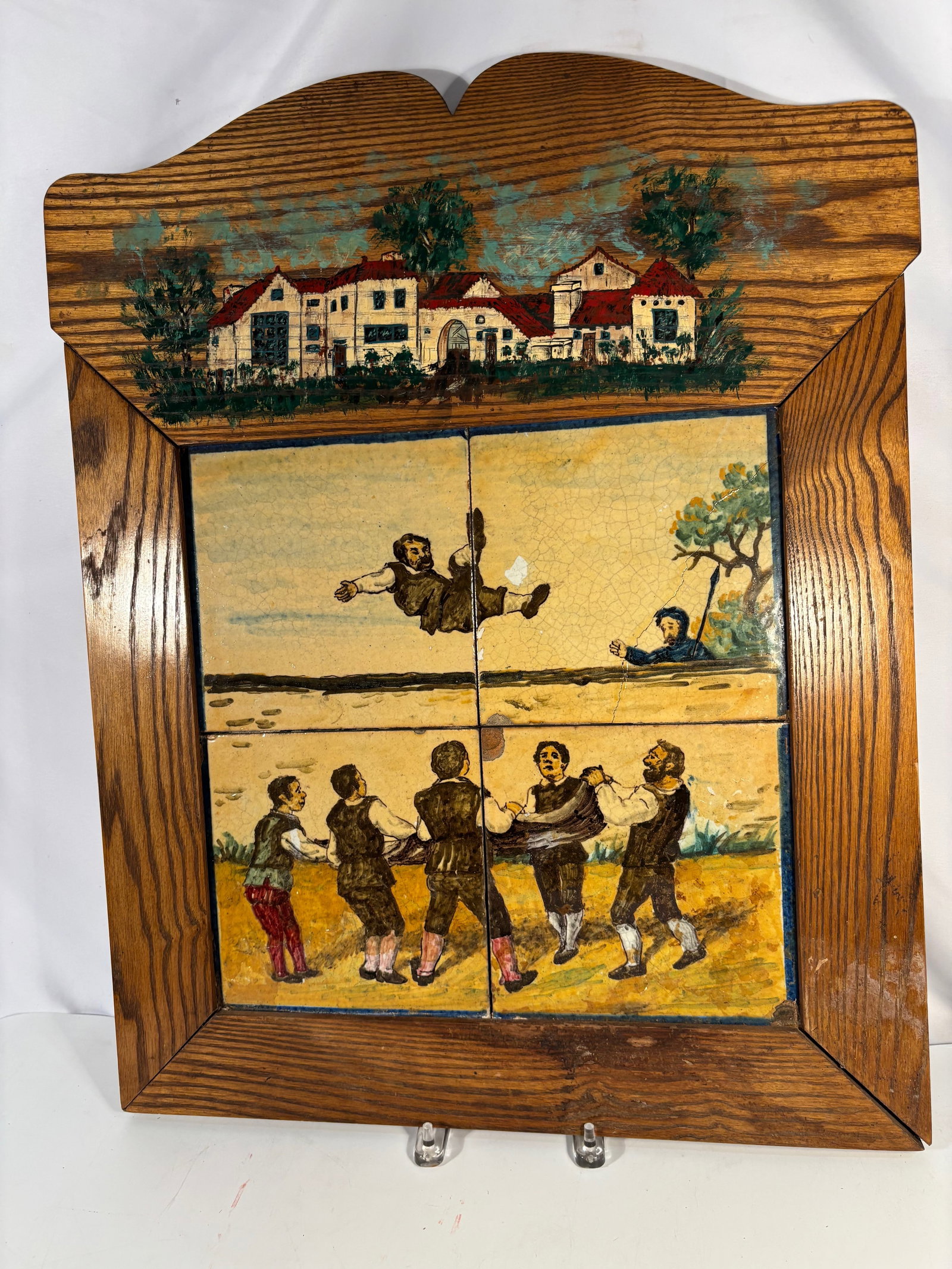 Antique European Hand Painted Four-Tile Ceramic Wall Plaque: Antique European hand-painted ceramic tile panel composed of four tiles depicting a rustic folk scene of villagers tossing a man in celebration. Mounted in a carved wooden frame with architectural dec