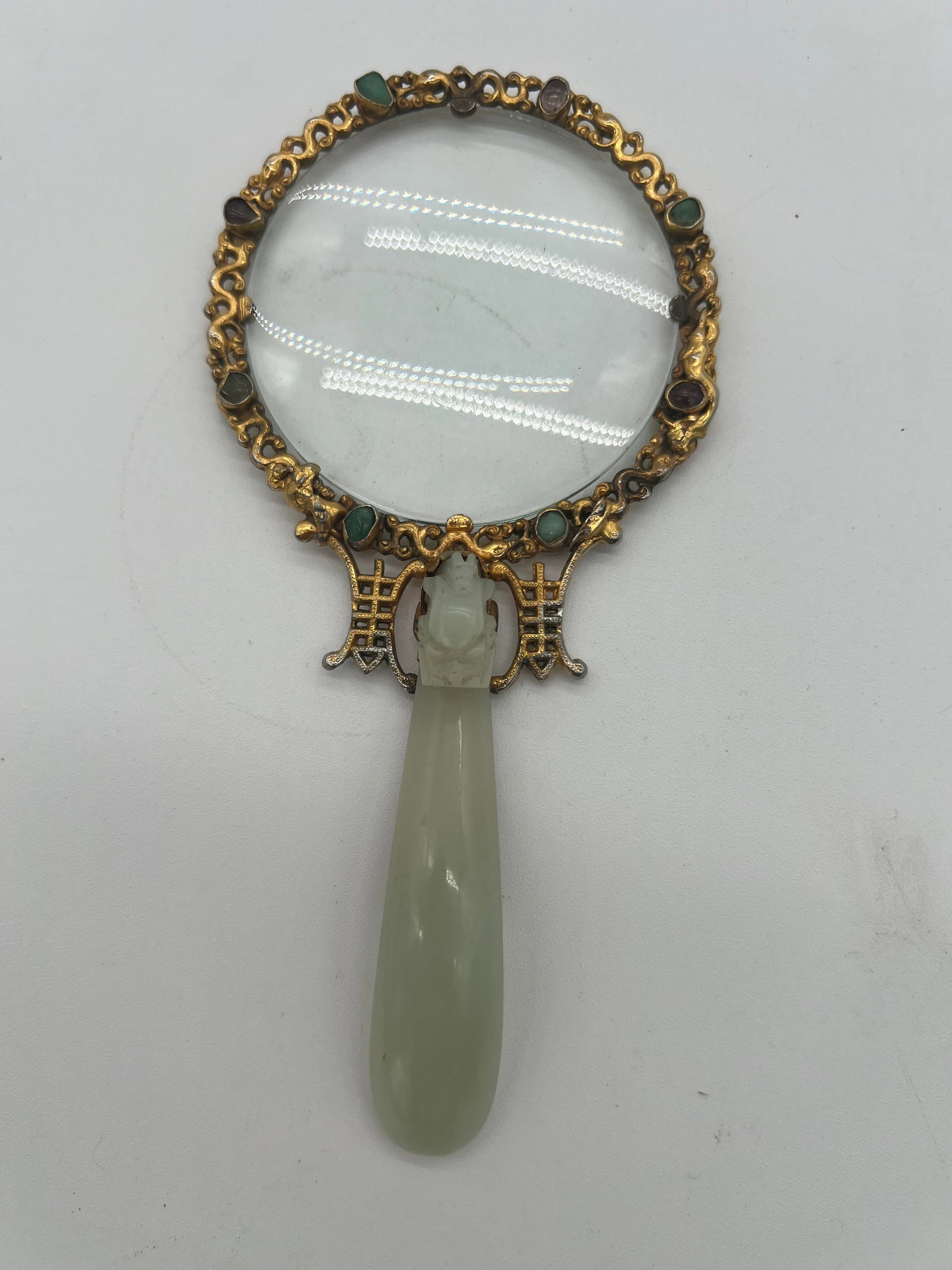 A gilt bronze framed magnifying glass with a jade handle and tourmaline gemstones: Qing Dynasty (1644-1912); The magnifying glass with a jade handle and is adorned 16 small gemstones like jadeite, tourmaline & etc. The frame surrounding the lens appears highly detailed, with a flora