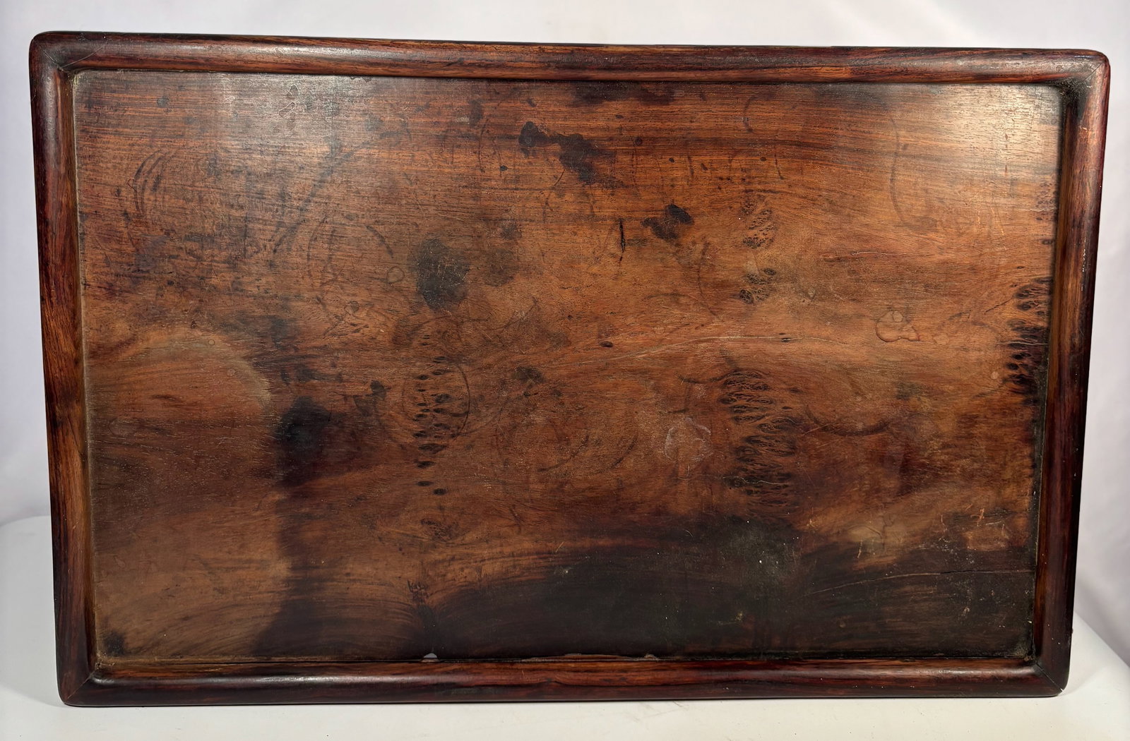 A Large Chinese Huanghuali Scholar’s Tray, Late Ming to Early Qing Dynasty: 17th–18th Century; A large rectangular huanghuali scholar’s tray, finely carved with raised edges and rounded inner lip, displaying a beautifully figured grain with swirling “ghost-face” p