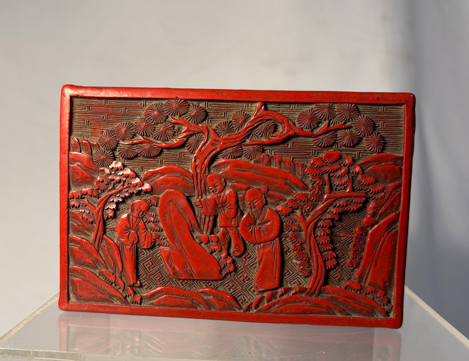 A Chinese Red Lacquer Carved Scholar’s Box: Early 20th century, A Chinese red lacquer style rectangular scholar’s box featuring finely carved figural and landscape decoration to the cover. The lid depicts robed figures in a mountainous garden