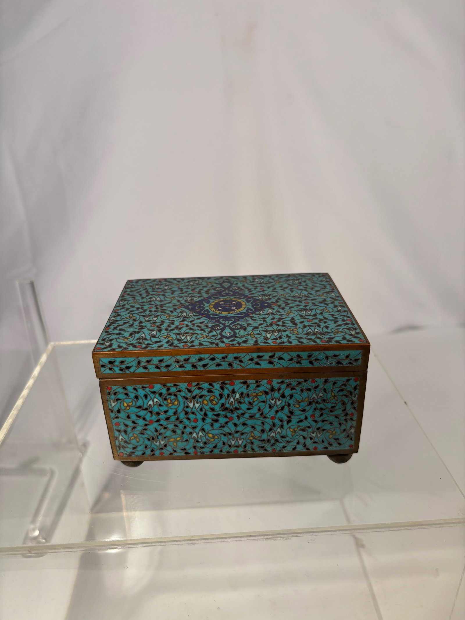 A Chinese Cloisonné Enamel Box, Qing dynasty(1644-1912): A finely crafted Chinese cloisonné enamel box, decorated with a dense turquoise-ground floral scroll pattern on all sides. The lid centered with a dark blue and gilt diamond-shaped medallion, compose