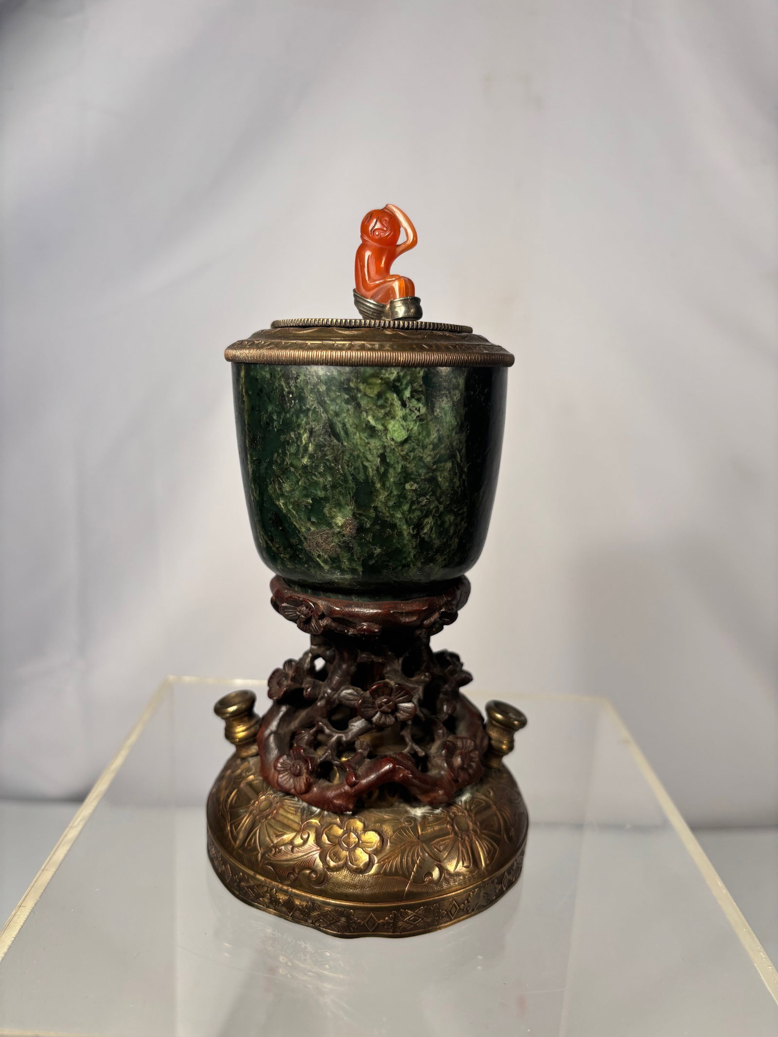 A Chinese Spinach Jade Inkwell with Gilt Bronze Stand and Cornelian Agate Monkey Finial: 19th century; A Chinese spinach jade inkwell of cup form, carved from dark green stone with natural mottling. The vessel is fitted with a gilt bronze cover surmounted by a finely carved cornelian agat