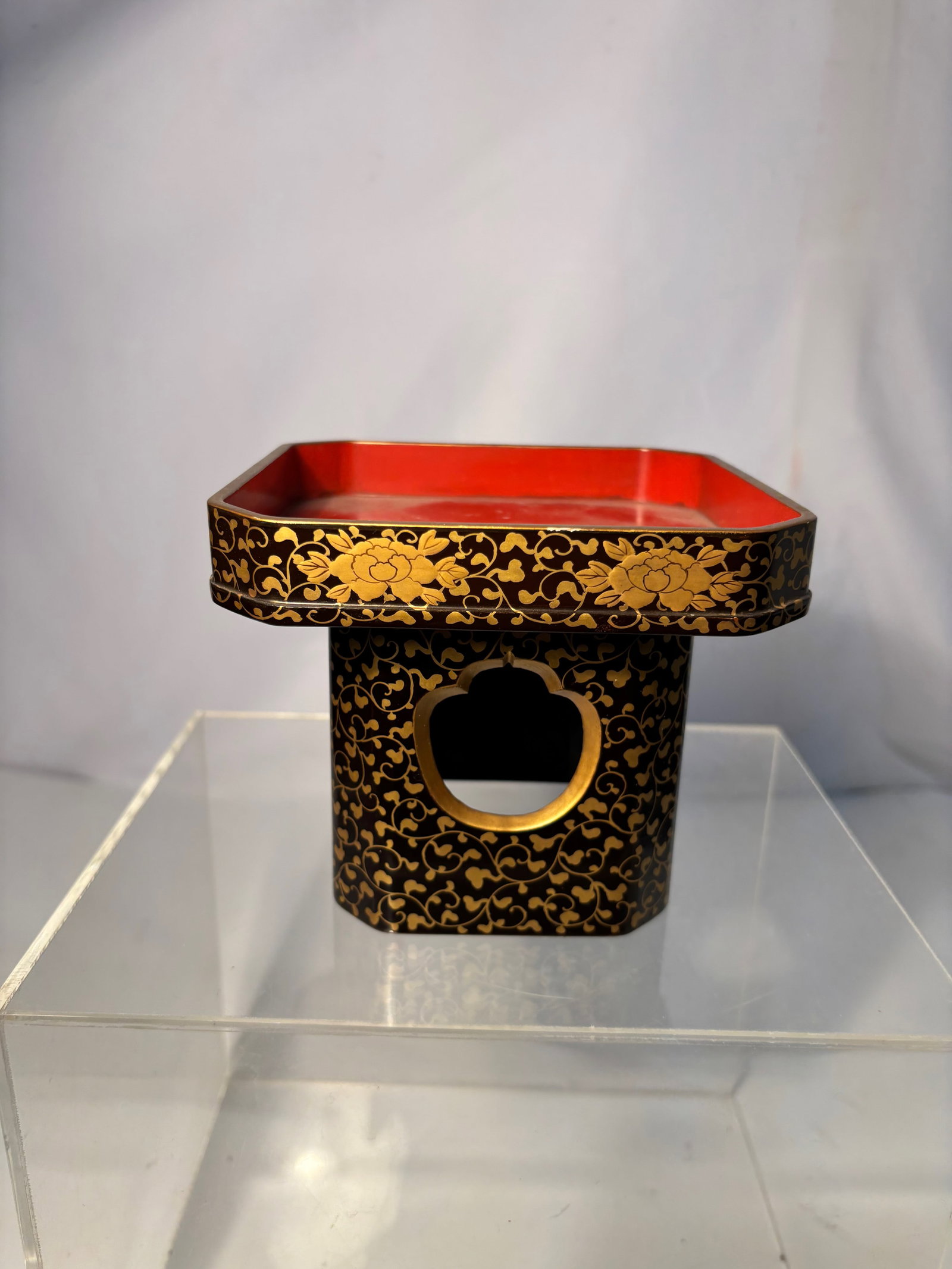 A Japanese Lacquered Wood Stand with Gilt Decoration, Figural Motifs: Japanese lacquered wood stand or small pedestal, finished in deep red and black lacquer with extensive gilt floral scrollwork. The sides are decorated with gilt figural scenes depicting mounted figure