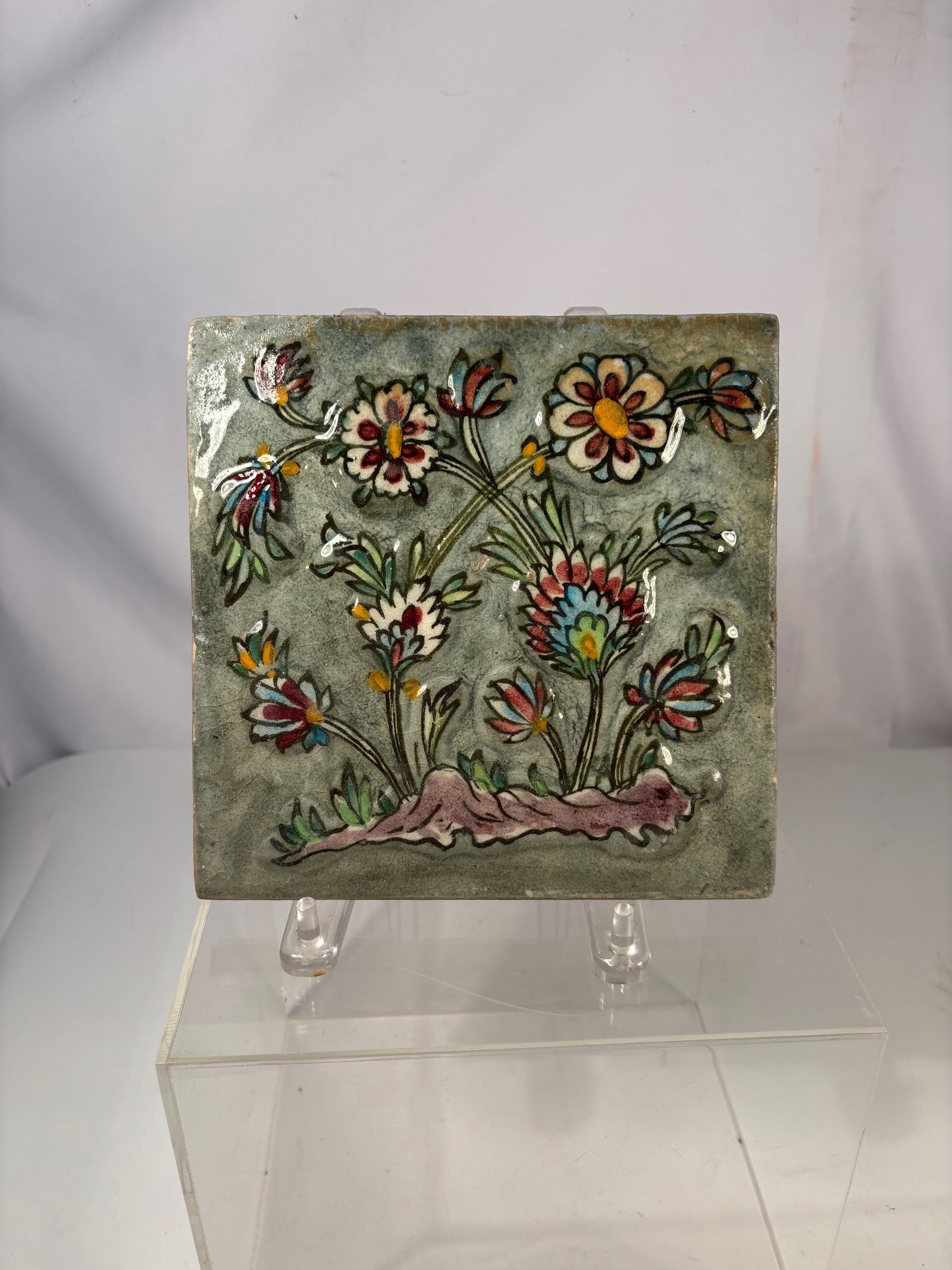 A Persian Qajar Polychrome Floral Glazed Ceramic Tile, 19th C.: A Persian Qajar period glazed ceramic tile decorated with a stylized floral spray in polychrome enamels on a grey-green ground. The design features flowering stems emerging from a mound, rendered in b