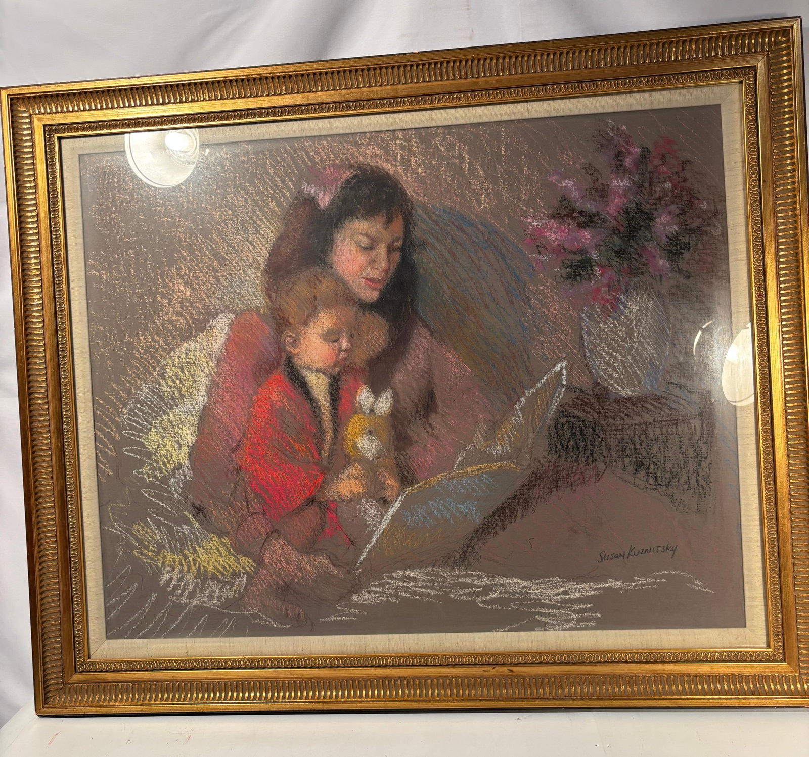 Susan Kuznitsky Signed Pastel, Mother and Child Reading Scene: A warm and beautifully rendered original pastel drawing by Susan Kuznitsky, depicting a mother reading with a young child seated in bed, holding a stuffed rabbit. The composition includes a floral arr