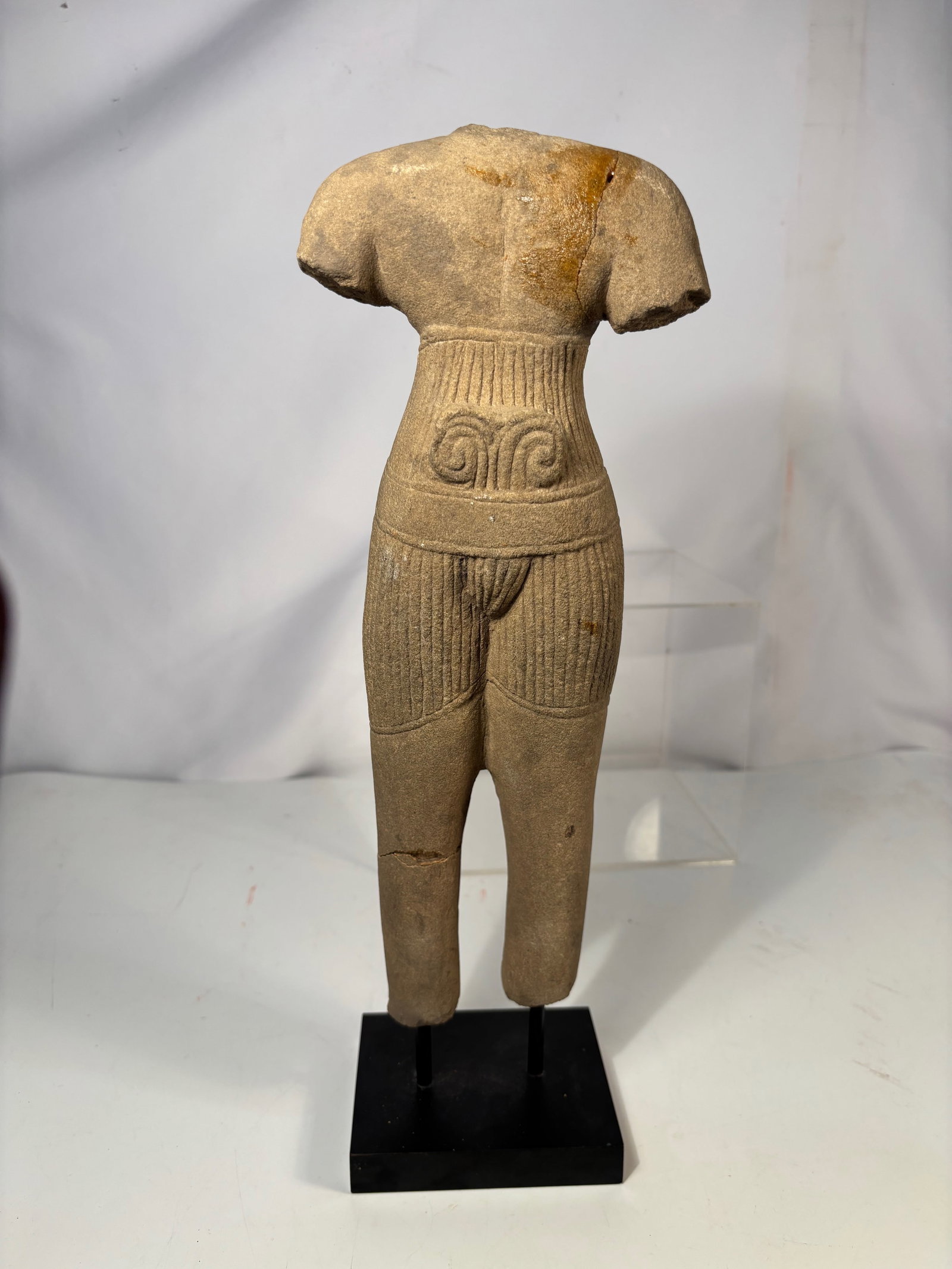 Khmer Style Carved Sandstone Male Deity Torso: Carved sandstone male deity torso in the style of Khmer Angkor sculpture. The figure displays incised garment folds and a decorated belt with scroll motifs. Fragmentary form without head and arms. Mou