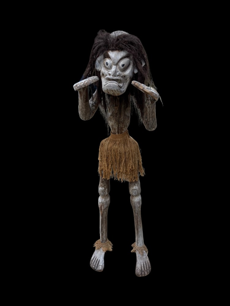 An African Tribal Carved Wood Figure with Fiber Hair and Raffia Skirt: Carved wooden tribal figure depicting a stylized human form with exaggerated facial features, including bulging eyes and a pronounced mouth. The figure is shown standing with arms raised near the face