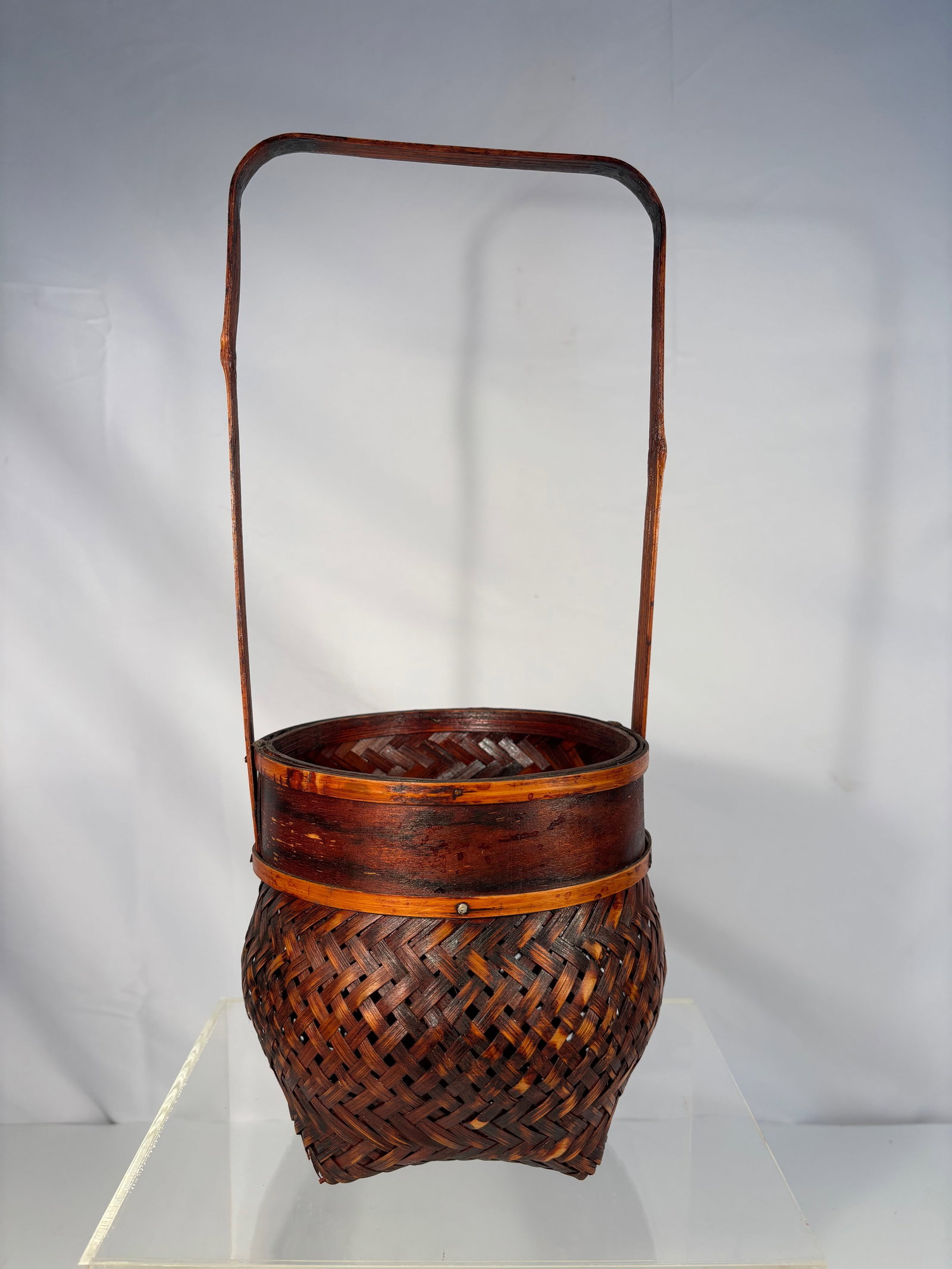 A Japanese Woven Bamboo Ikebana Flower Basket with Tall Handle: A finely handwoven bamboo basket with tall fixed handle, likely Japanese, early 20th century. The basket body displays a tight diagonal plait weave with warm reddish-brown lacquered patina. The rim is