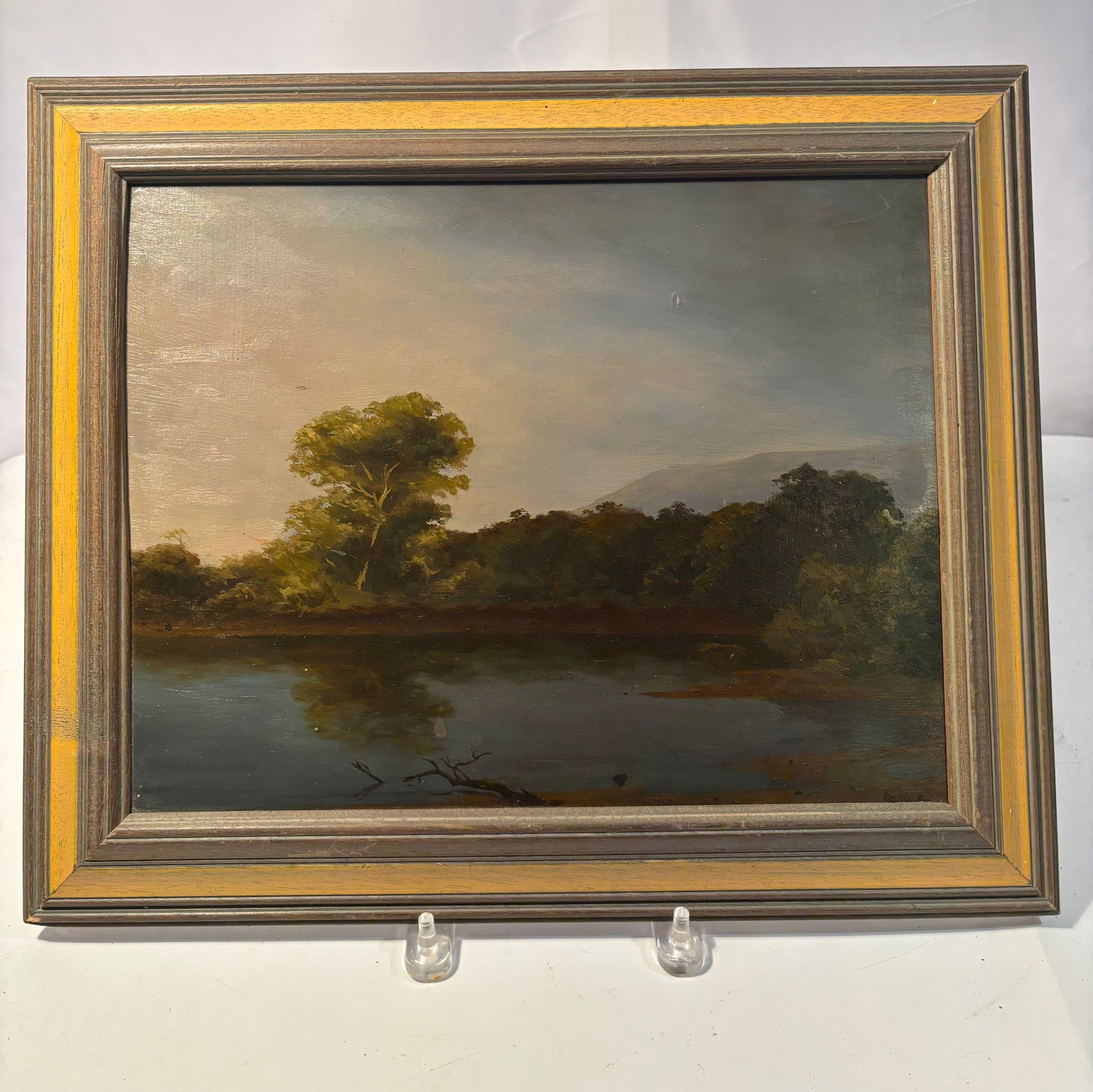 Framed Landscape Oil Painting, Signed Patricia Berger, 1993: This piece features a tranquil landscape scene depicting a calm body of water bordered by trees, with soft atmospheric light and gentle reflections across the surface. The composition centers on a sol
