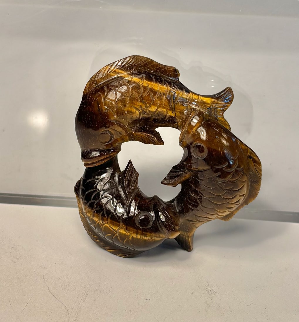 A Tiger’s Eye Stone Carving of Three Fish in Circular Form: A finely carved circular sculpture depicting three intertwined fish, rendered in richly colored tiger’s eye stone. The piece displays beautiful natural chatoyancy, with golden and brown bands shifti