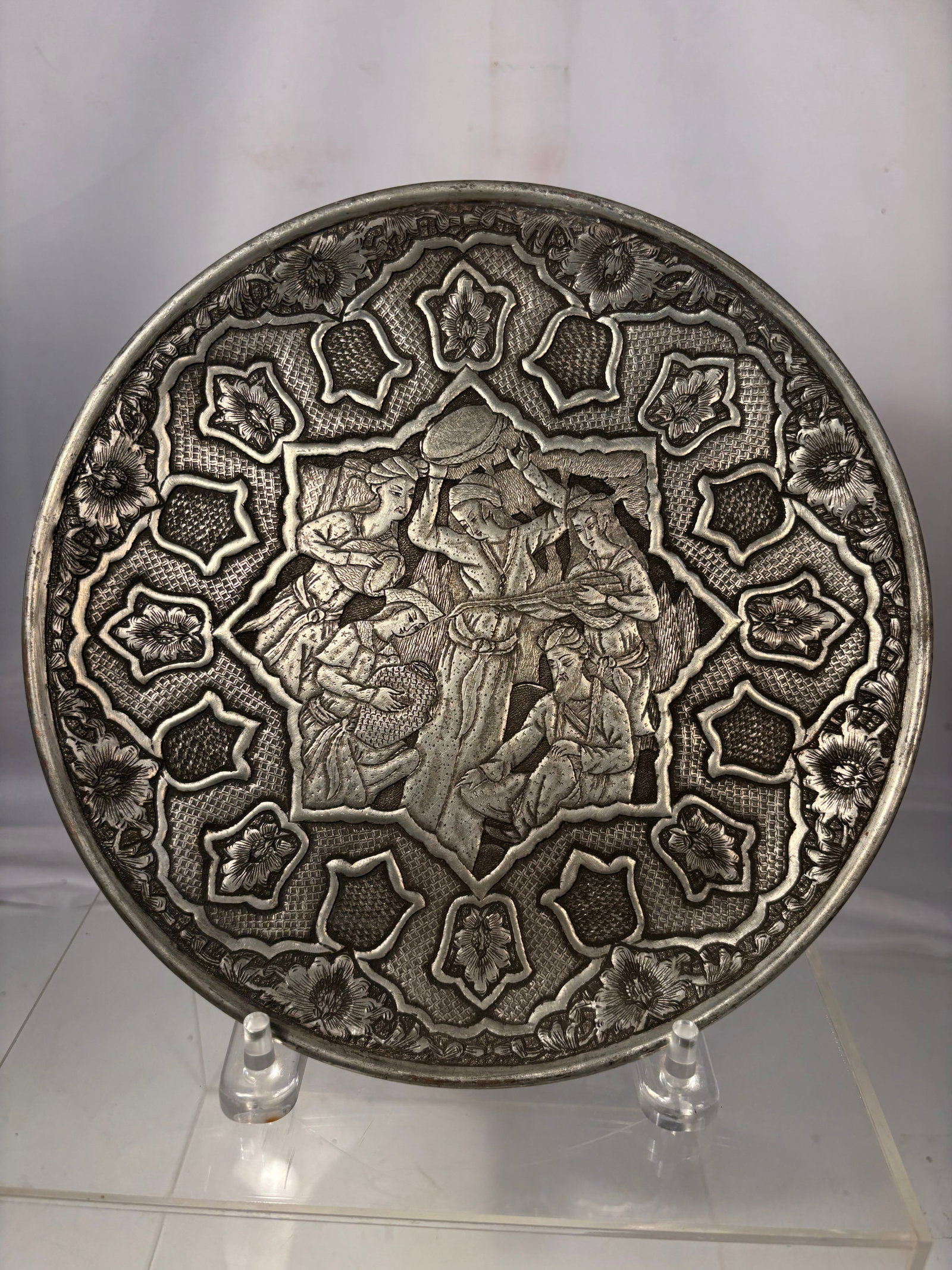 Hand-Engraved Persian Isfahan Ghalamzani Silvered Copper Decorative Charger: Persian-style circular decorative plate featuring a central figural scene surrounded by geometric and floral motifs in relief. The design depicts several figures in traditional attire within a shaped 