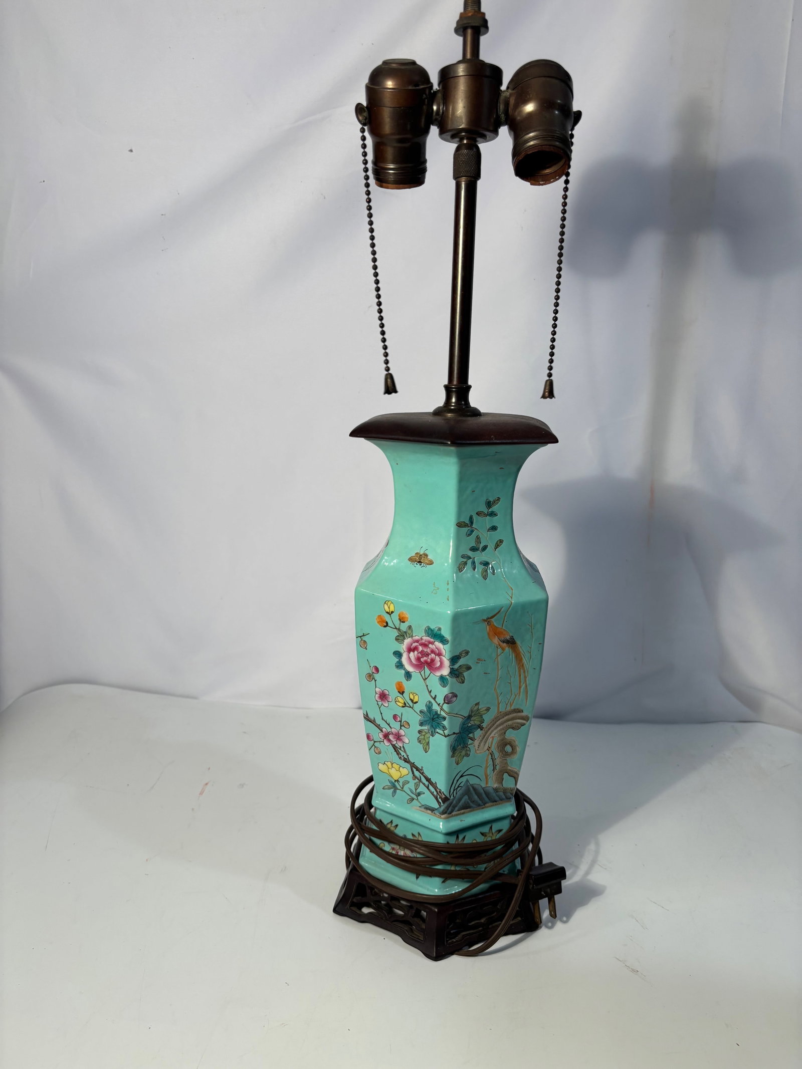 A Chinese Turquoise Ground Famille Rose Porcelain Vase Mounted as a Table Lamp , 19th century: A Chinese porcelain vase decorated with famille rose enamels on a turquoise ground, featuring birds, flowers, and insects in a garden setting. The faceted body rising from a fitted wooden stand. Later