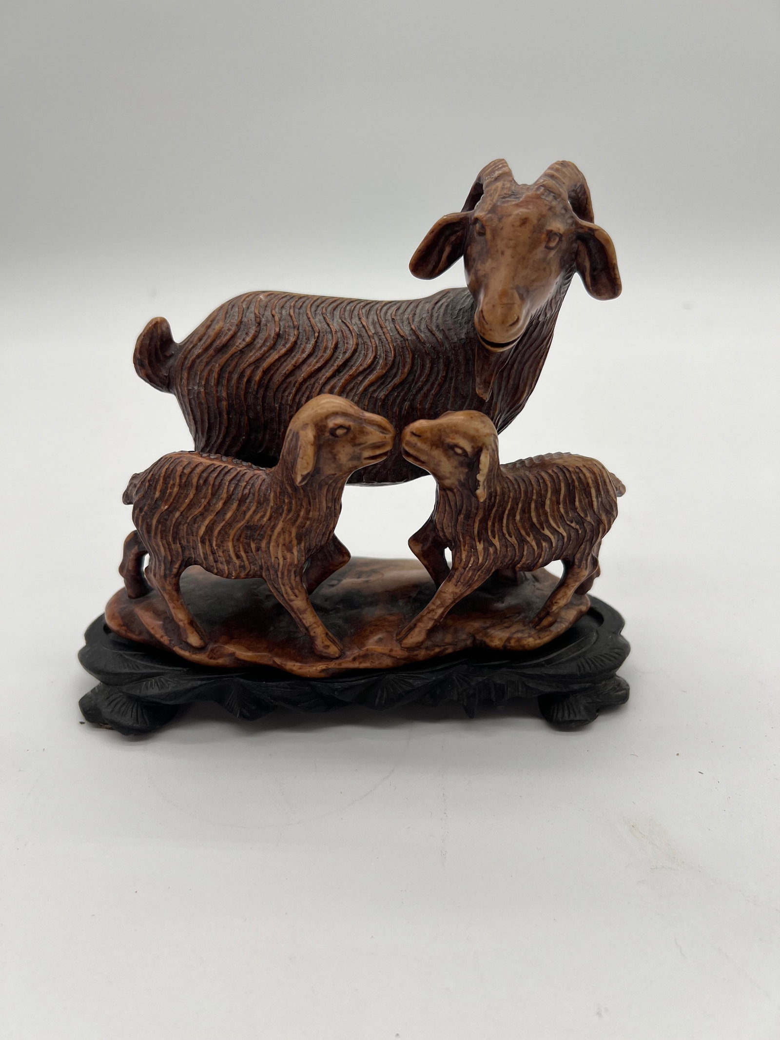 A Chinese hand carved three goats statue with wood base: Late 19th or early 20th; This family of goats or sheep carved in soapstone likely represents a symbol of harmony and nurturing, which is often portrayed in pastoral scenes or animal-themed carvings. S