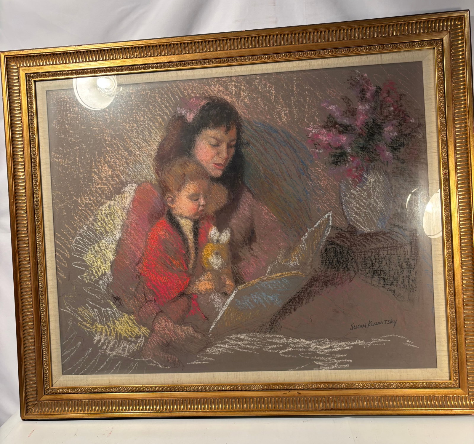Susan Kuznitsky Signed Pastel, Mother and Child Reading Scene: A warm and beautifully rendered original pastel drawing by Susan Kuznitsky, depicting a mother reading with a young child seated in bed, holding a stuffed rabbit. The composition includes a floral arr