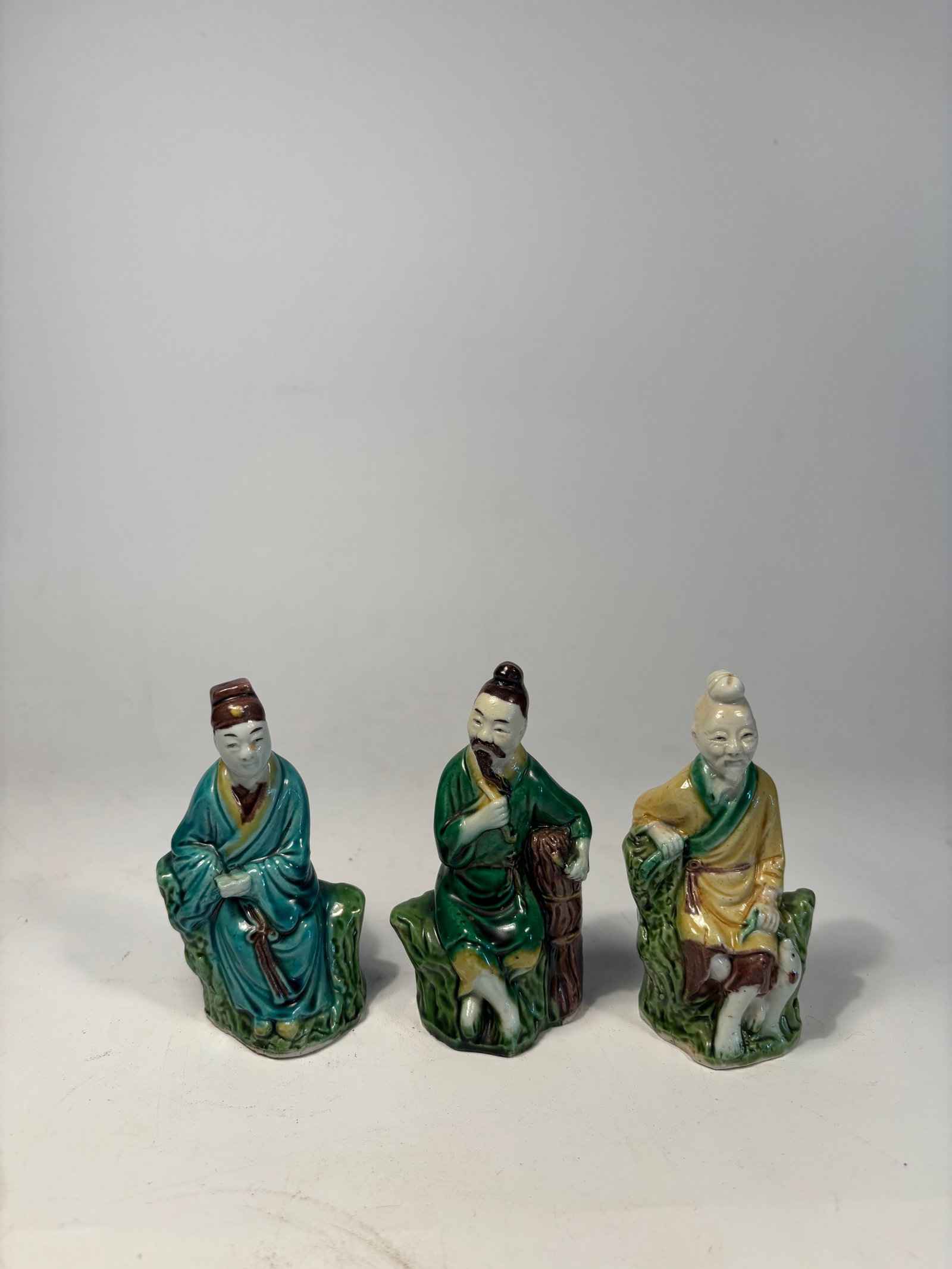Set of Three Chinese Sancai-Glazed Porcelain Scholar Figures: Early 20th century; A set of three seated Chinese porcelain scholar figures, each finely modeled and decorated in traditional sancai glaze tones of green, amber, and cream. Each figure is portrayed in
