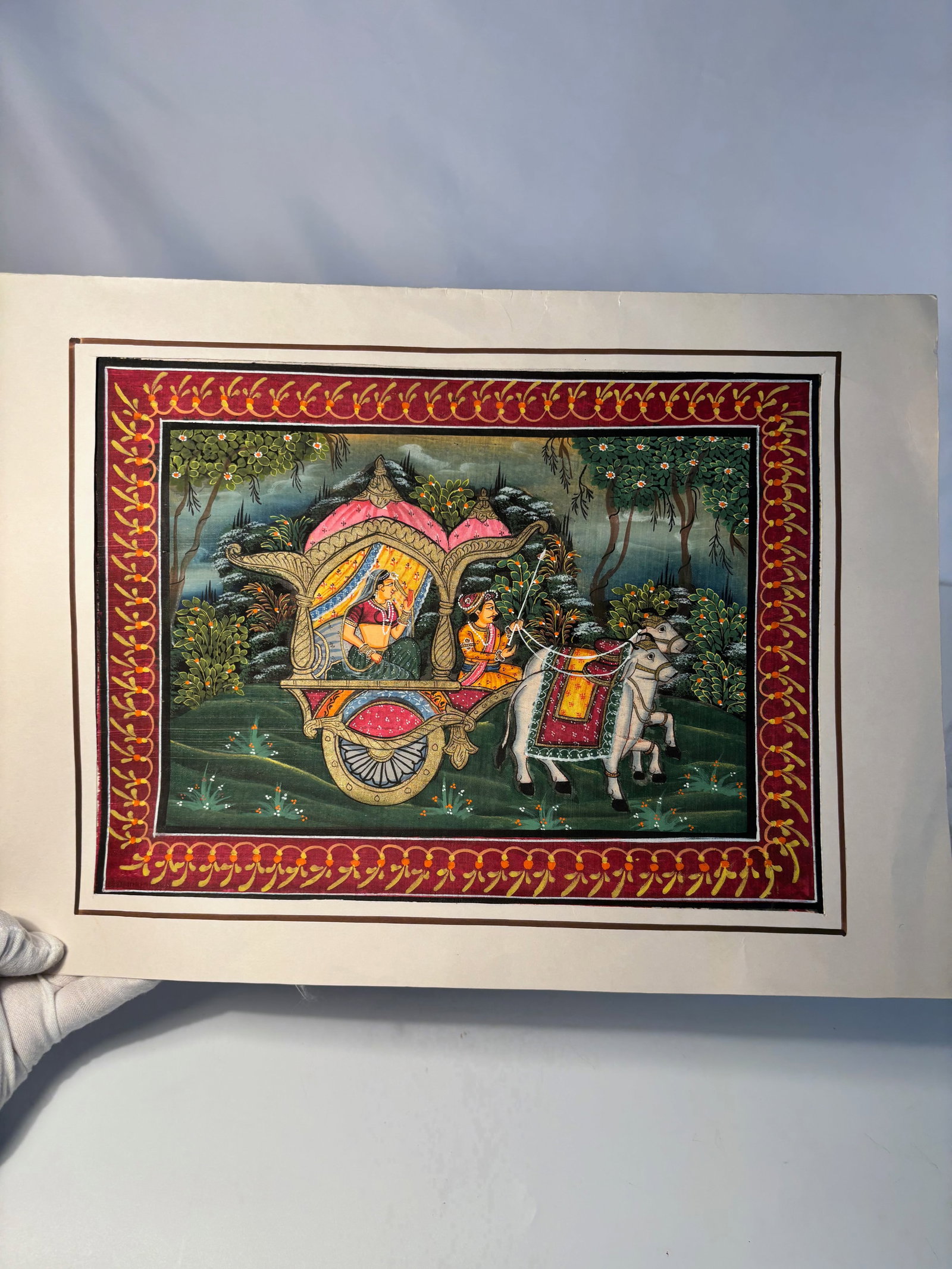 Indian Miniature Gouache on Paper, Noble Couple in Ox-Drawn Chariot: 19th century to 20th century; A hand-painted Indian miniature in gouache on paper, depicting a richly adorned nobleman and lady riding in an ox-drawn chariot through a verdant landscape. The chariot i