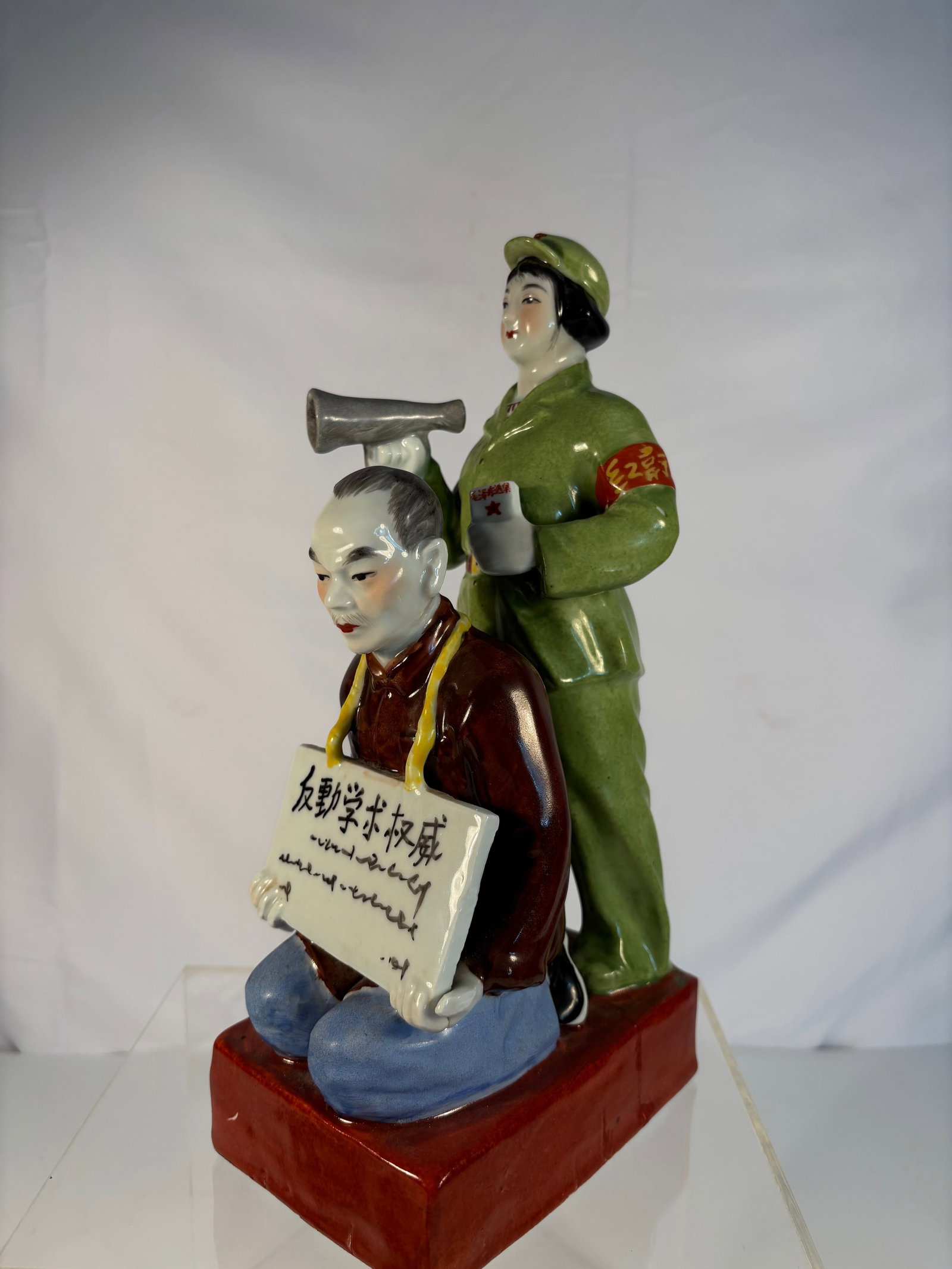 A Chinese Cultural Revolution Porcelain Figures statue: Cultural Revolution (1966–1976); A woman in a green Red Guard uniform is holding a loudspeaker and a book (likely Mao’s Little Red Book). She stands behind a kneeling man wearing a placard. Dimens