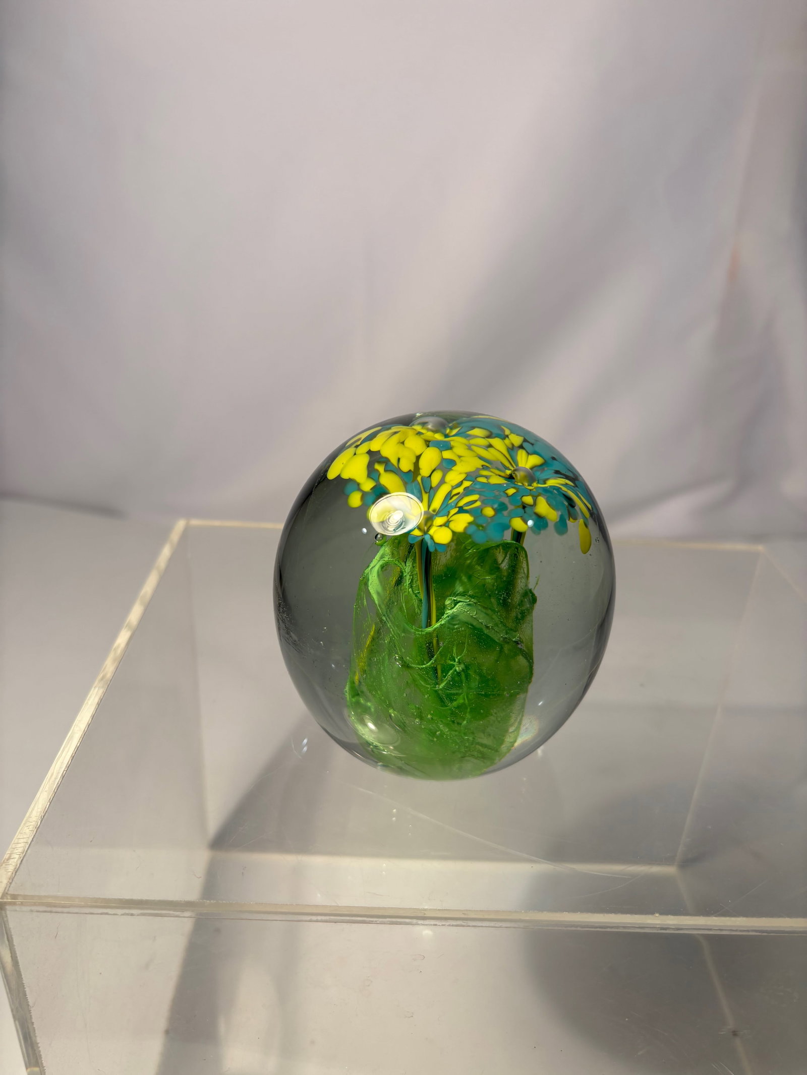 A Kerry Glass Art Glass Floral Paperweight, Hand Made in Ireland: Art glass paperweight by Kerry Glass, hand made in Ireland. The spherical form encloses a stylized floral motif composed of green, yellow, and blue glass elements, suspended within a clear glass body.