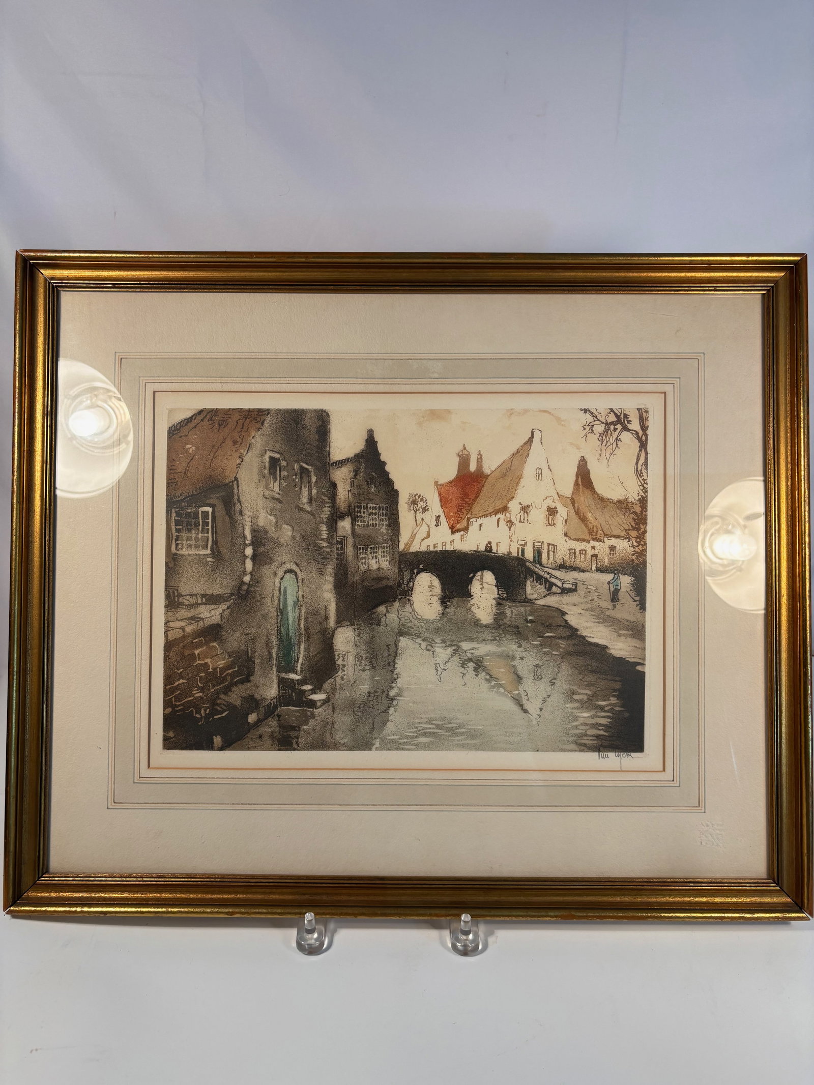 Van Lyck (European, Early 20th Century) — Hand-Colored Etching of a Canal Town with Bridge: Depicting a European canal town with steep-gabled houses, a stone bridge, and shimmering water reflections. Executed as an etching with aquatint, delicately hand-colored in soft tones of red, ochre, a