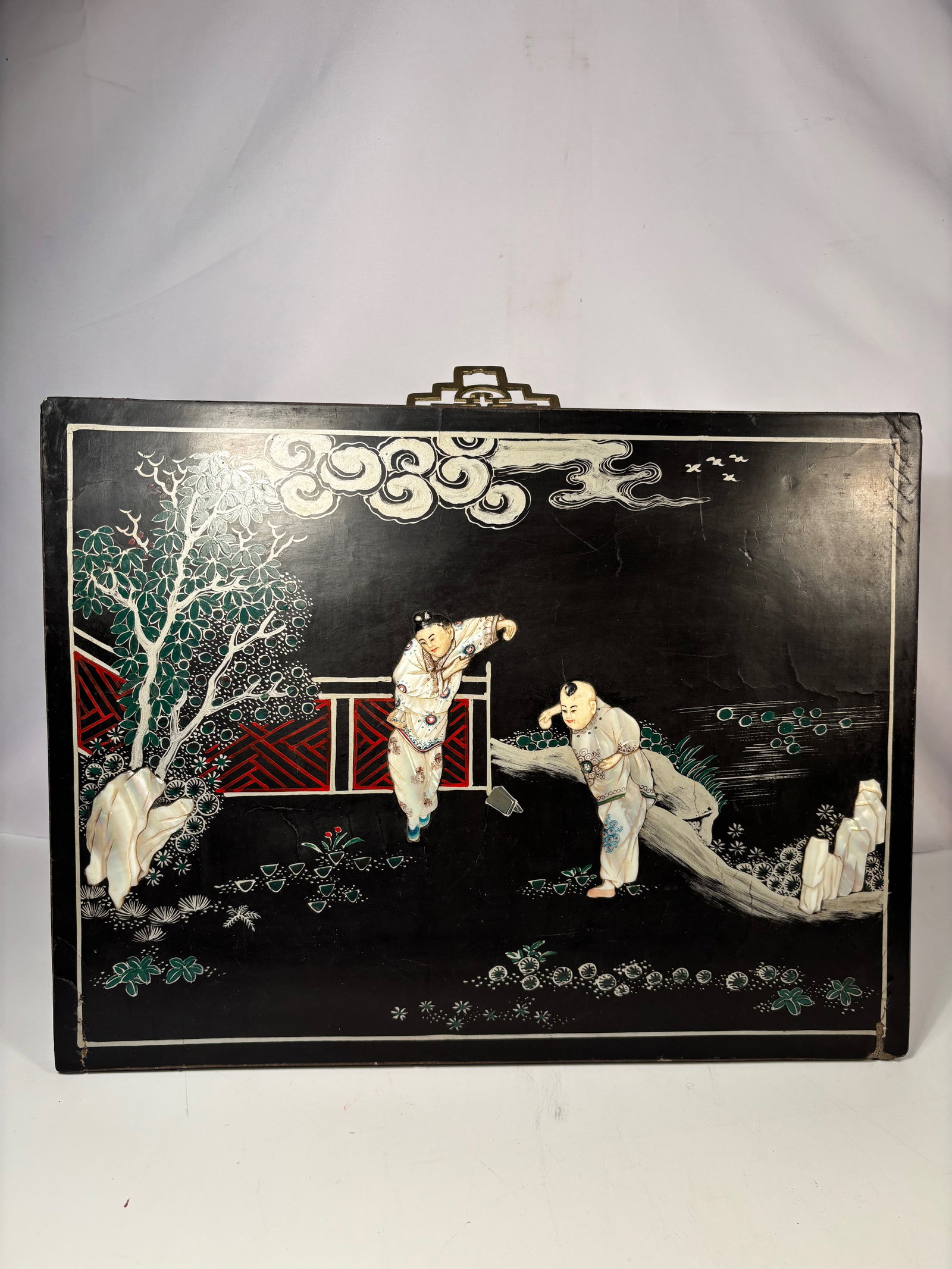 A Chinese Black Lacquer Mother-of-Pearl Inlaid Panel: Rectangular Chinese lacquer panel decorated with finely cut mother-of-pearl inlay depicting a figural garden scene. Two figures are shown in animated poses within a landscaped setting featuring rocks,