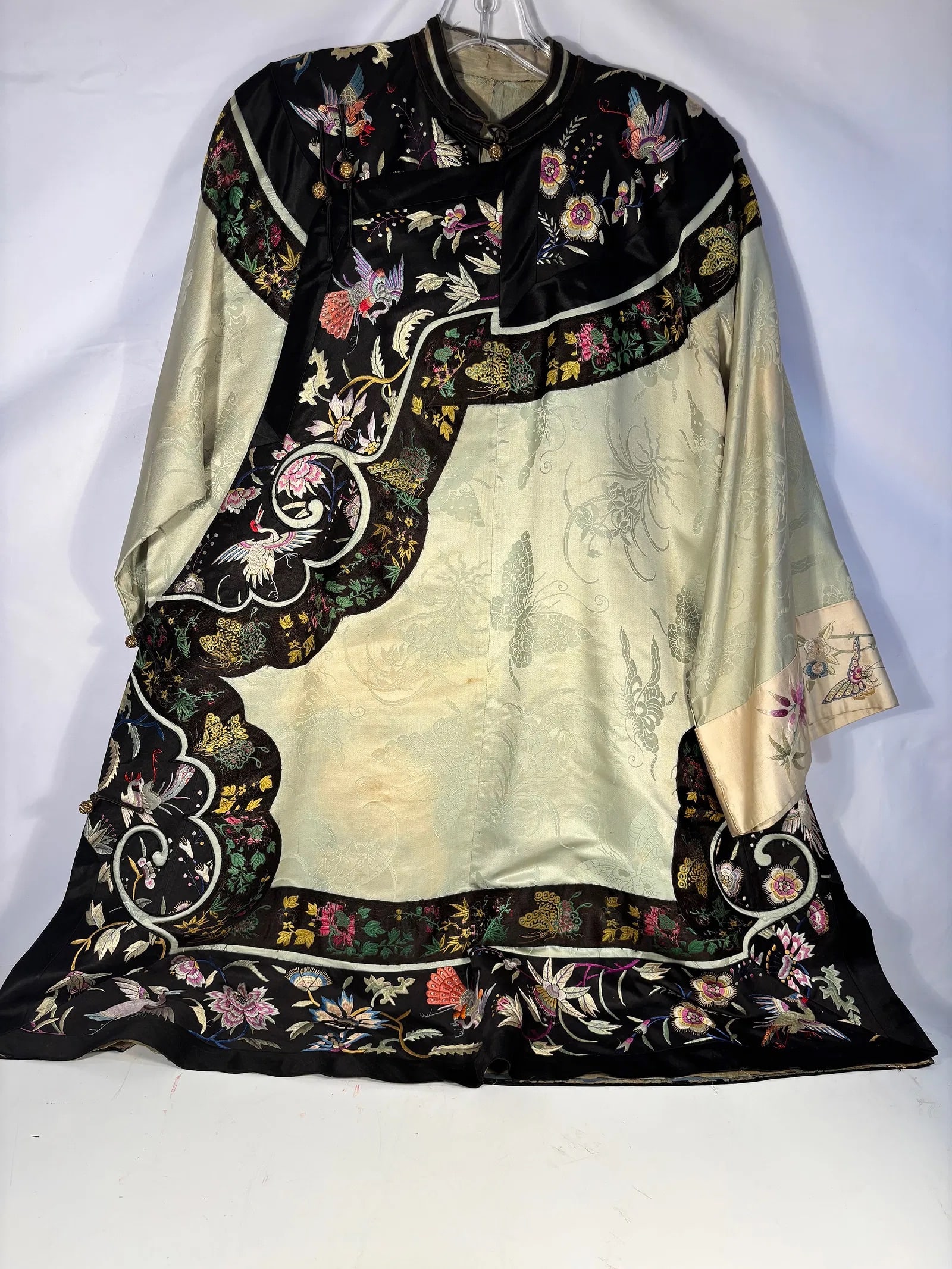 A Chinese Embroidered Silk Jacket: Chinese silk jacket, finely embroidered with cranes, butterflies, flowering branches, and auspicious motifs. The garment features a black satin ground with contrasting pale celadon-toned silk panels w