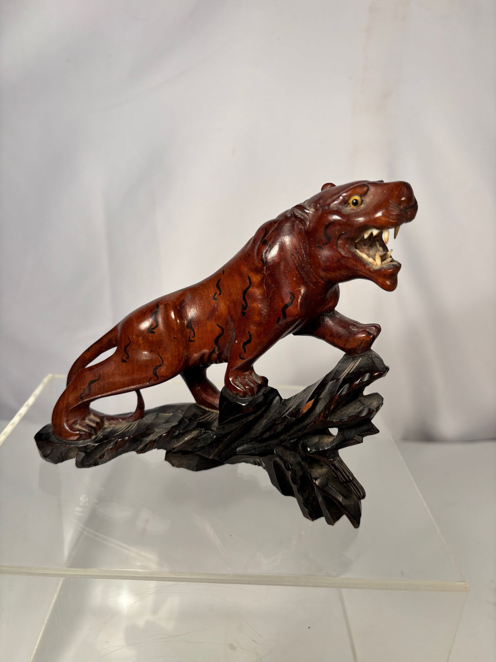 A Japanese Style Carved Tiger Figure on Wood Base: A Japanese-style carved figure depicting a tiger in a dynamic stalking pose, mounted on a carved base. The animal is rendered with expressive detail, including open jaws, incised striping, and inlaid-